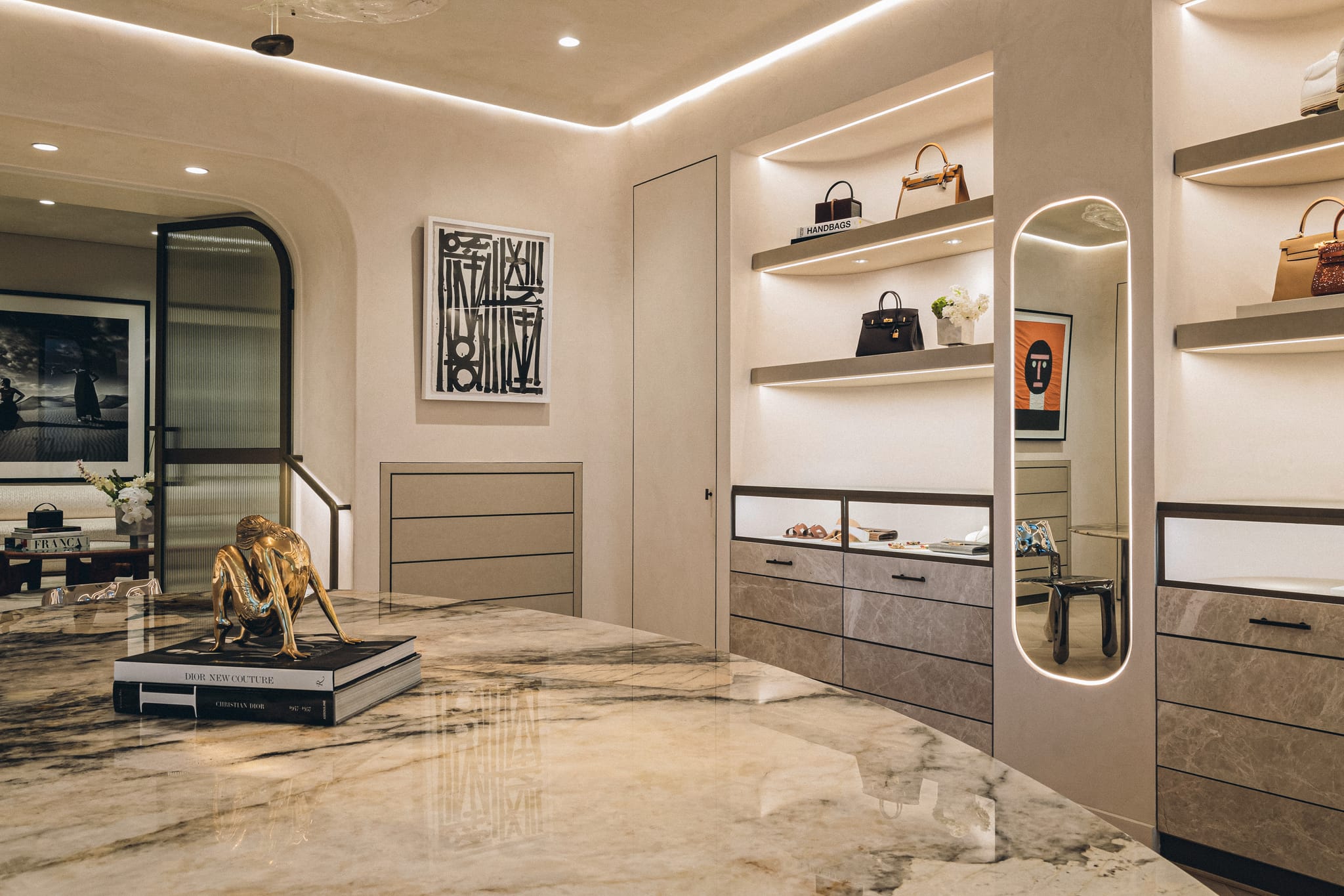 Veined marble display table holding a gilt figurine on art books, beside lit shelves of handbags and a brass-framed arched mirror.