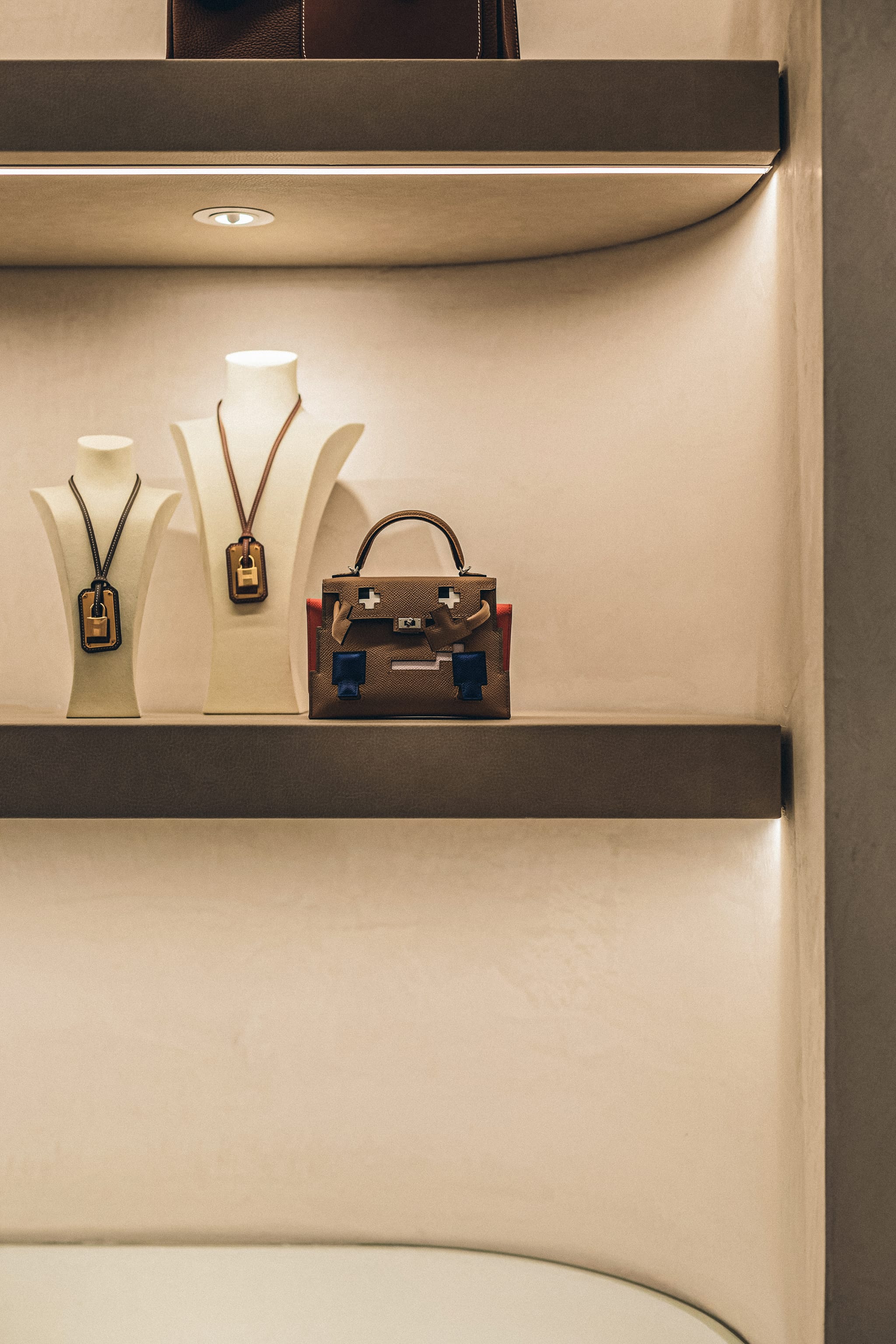 Close-up of leather pendant necklaces on cream busts and a small structured handbag on a brushed shelf under recessed lighting.