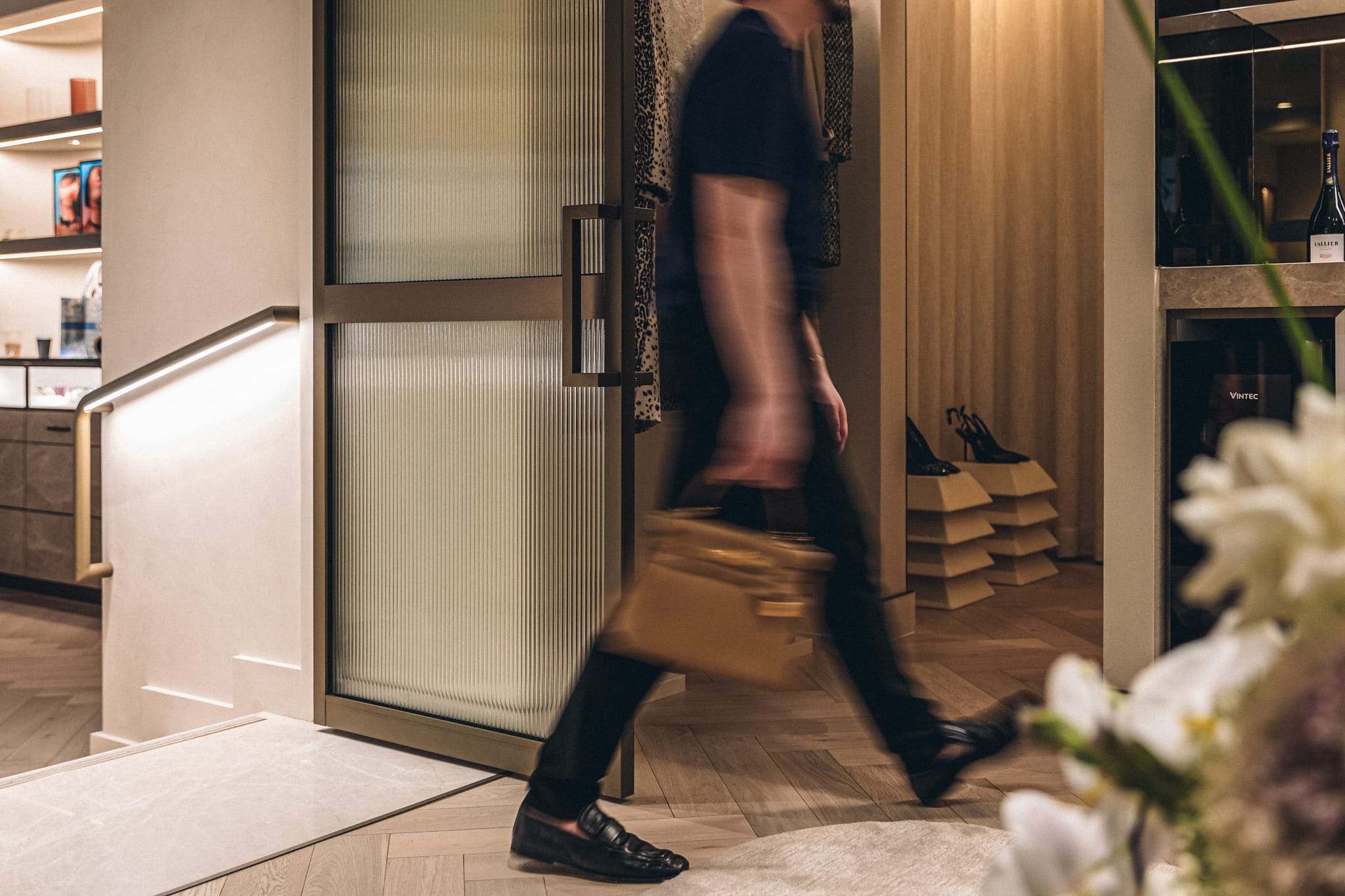 A figure moves past a reeded glass door carrying a case, with heels on stepped plinths and a wine cabinet in the retail interior.