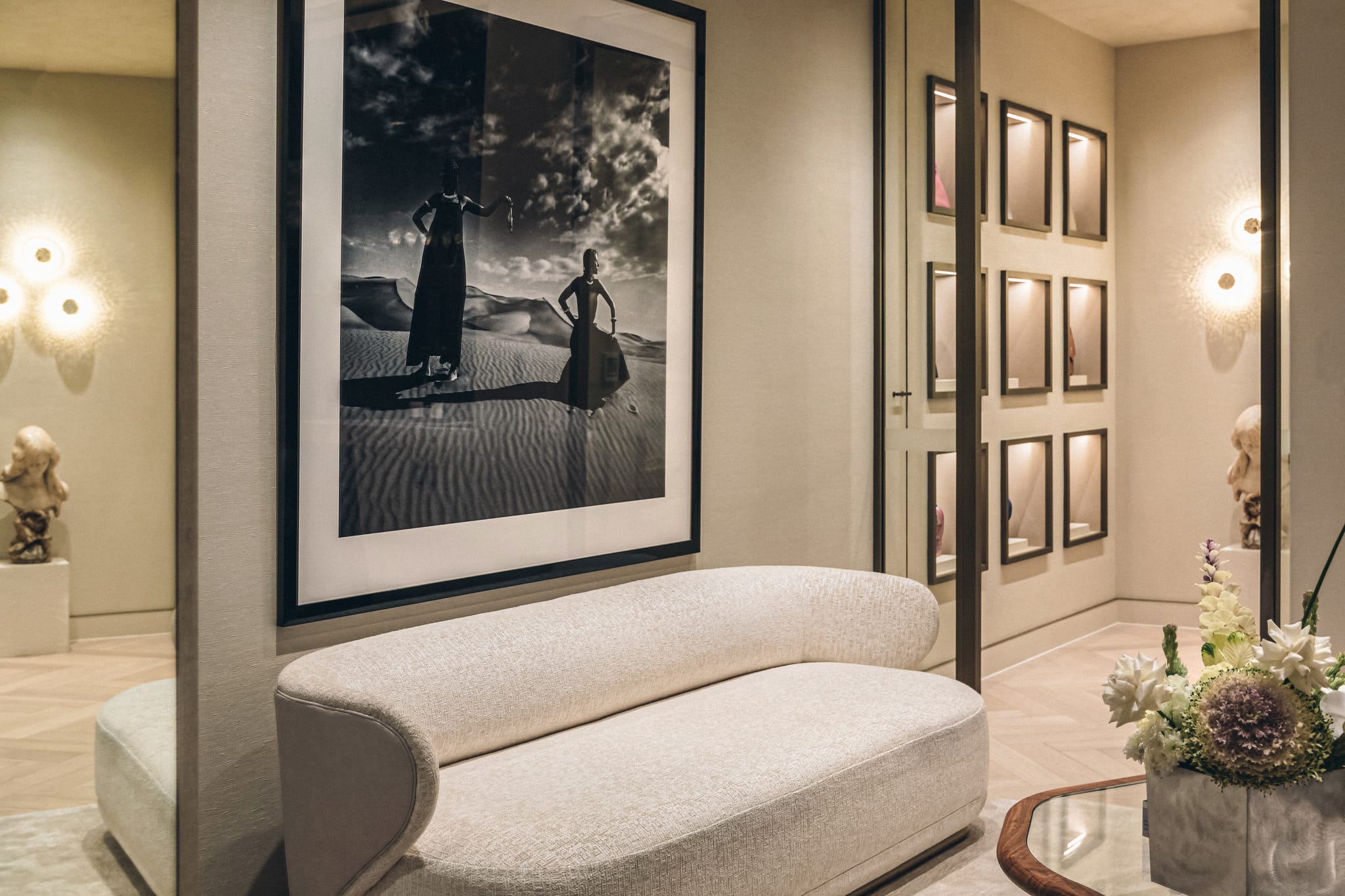 Curved cream bouclé sofa beneath a large black-and-white desert photograph, next to lit display niches and glass wall sconces.