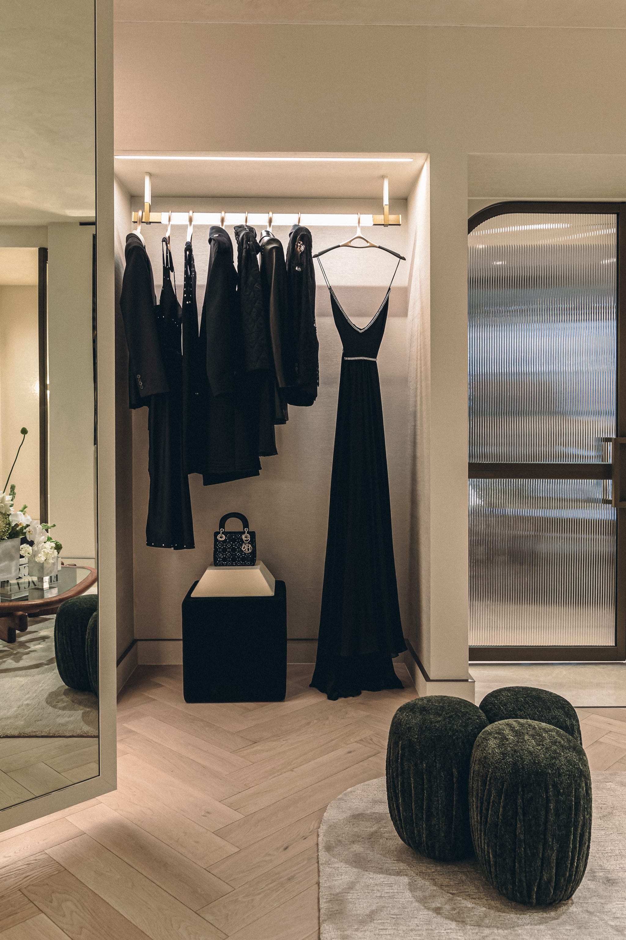 Black garments and an evening gown hang in a lit niche above a handbag on a plinth, with green velvet poufs and a reeded glass door.