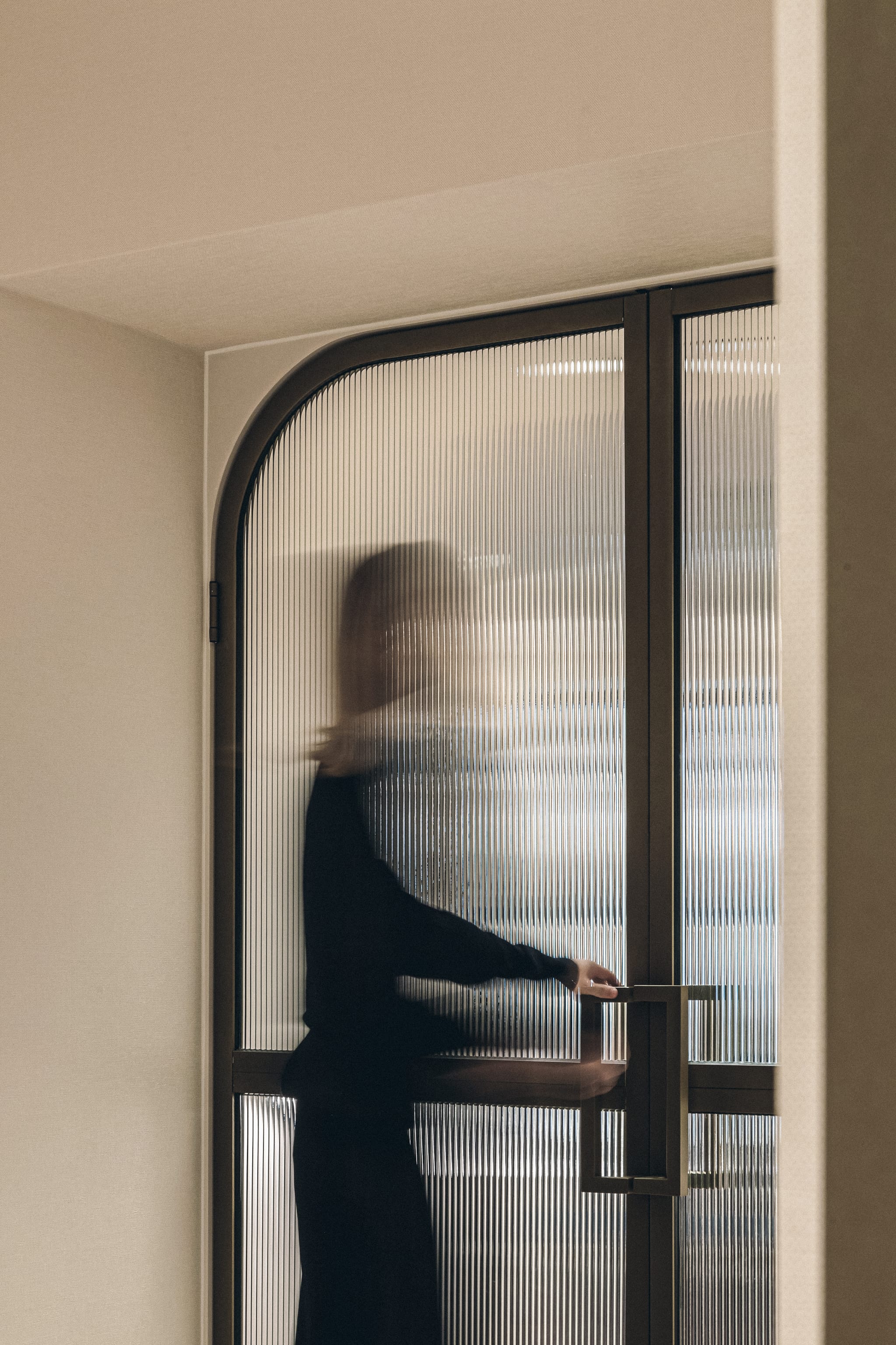 A figure in black seen through an arched reeded-glass door with a slim brass handle, softened by motion in the boutique fitting area.