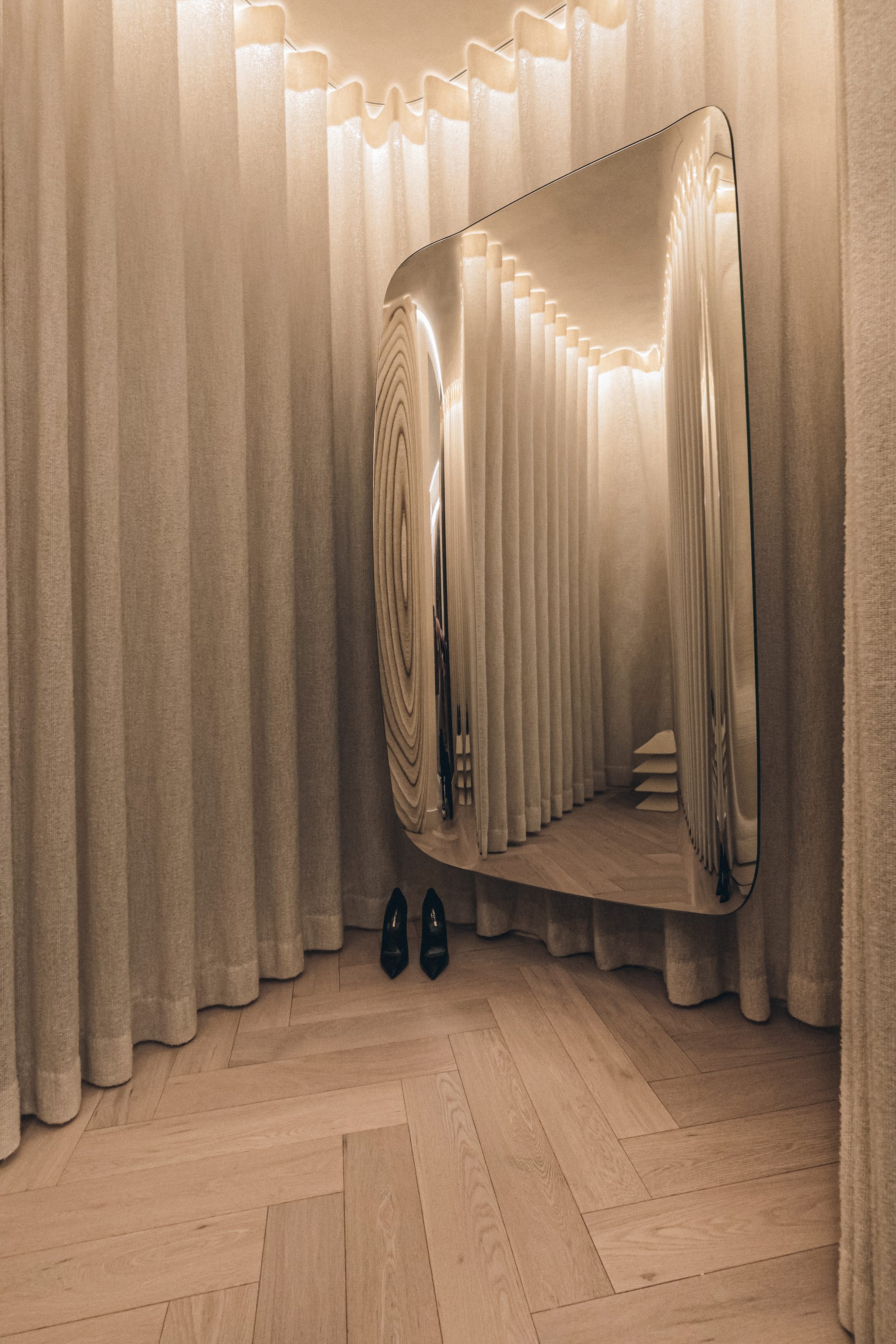 Fitting room wrapped in floor-length linen curtains, with a large curved distortion mirror and black heels on herringbone oak flooring.