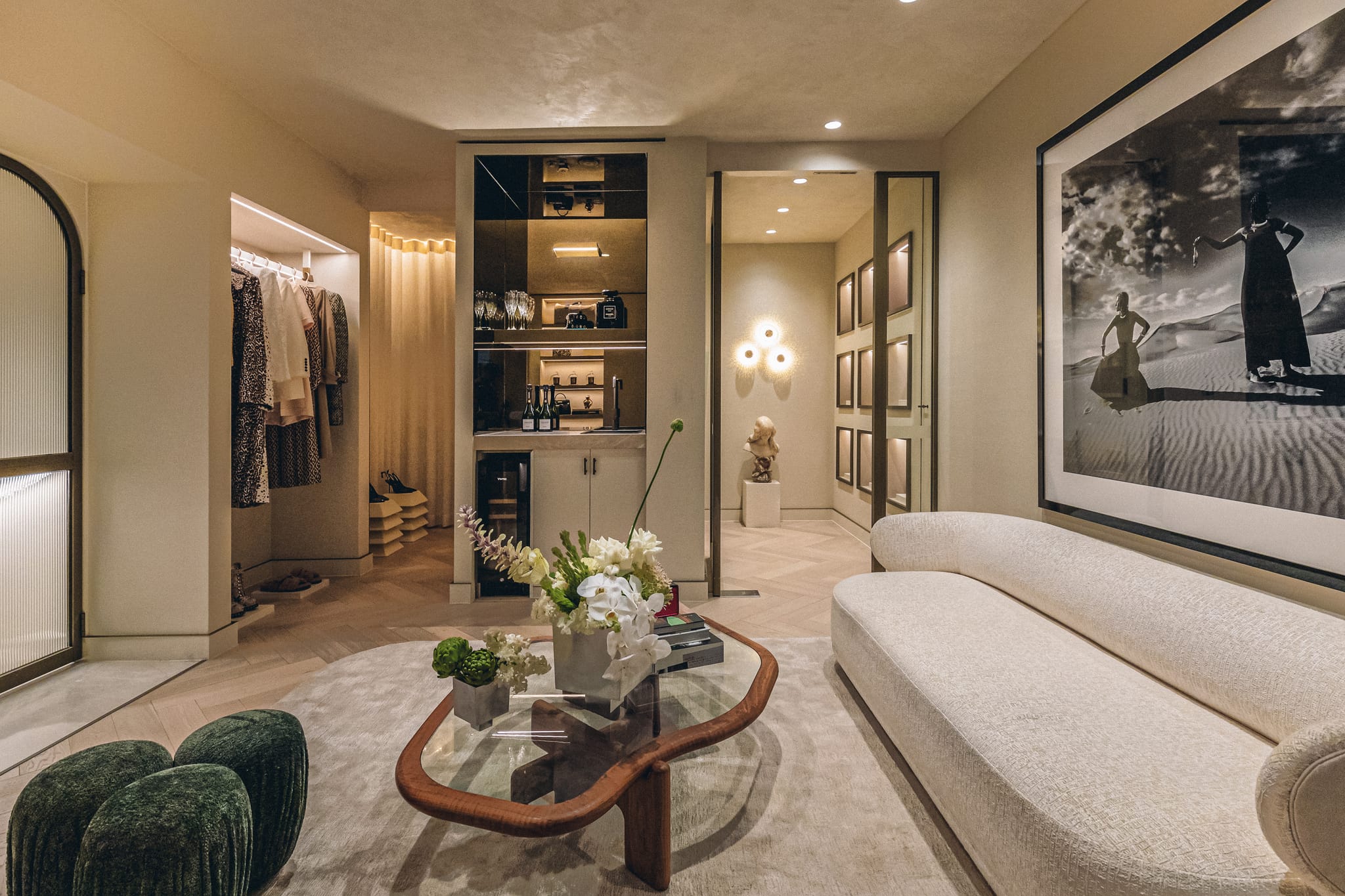 Lounge with a curved sofa, organic glass coffee table and floral arrangement, opening to a drinks bar and wardrobe in the retail interior.