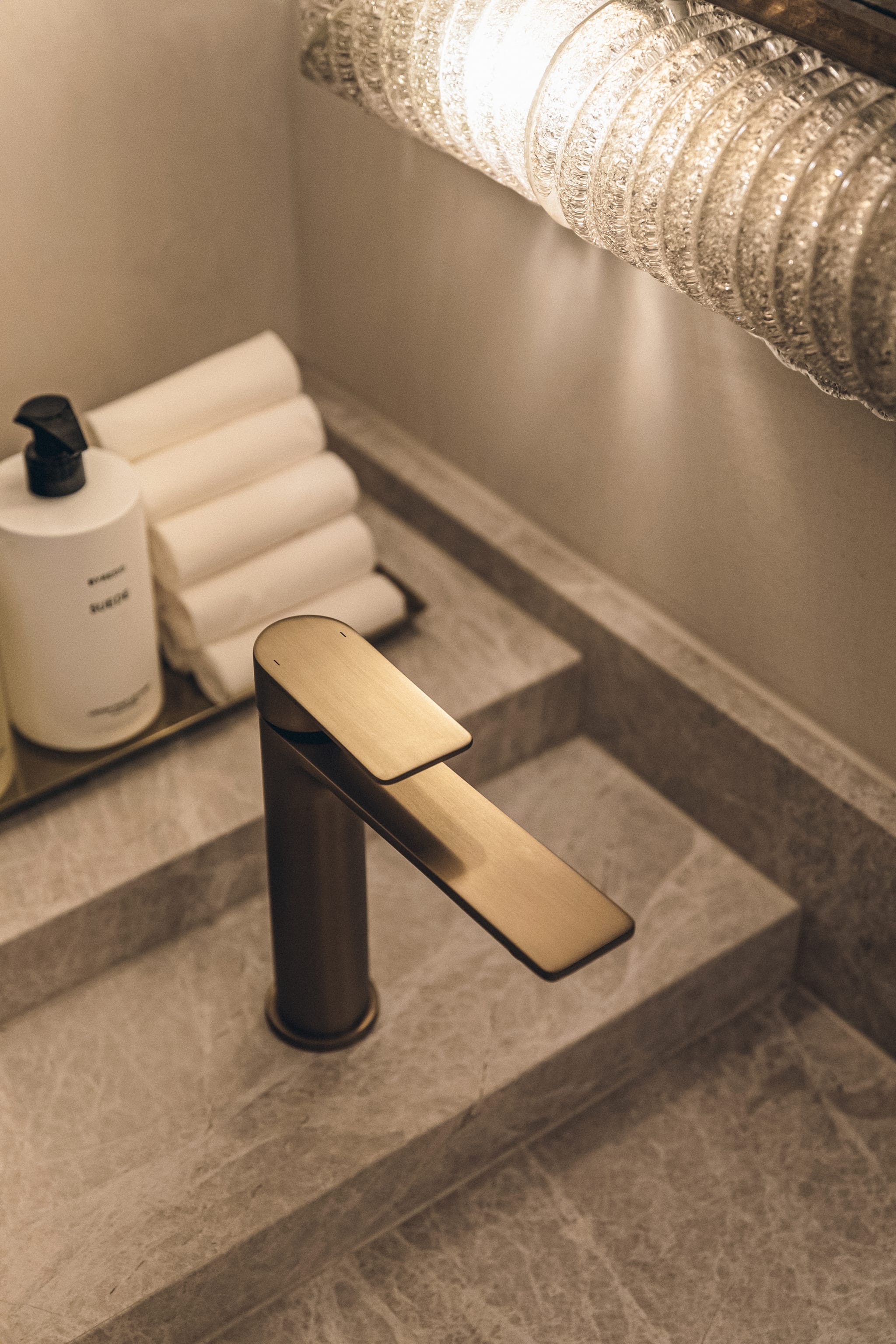 Brushed brass mixer tap on a grey marble vanity, with rolled white towels, a soap pump and a textured glass wall light in the washroom.