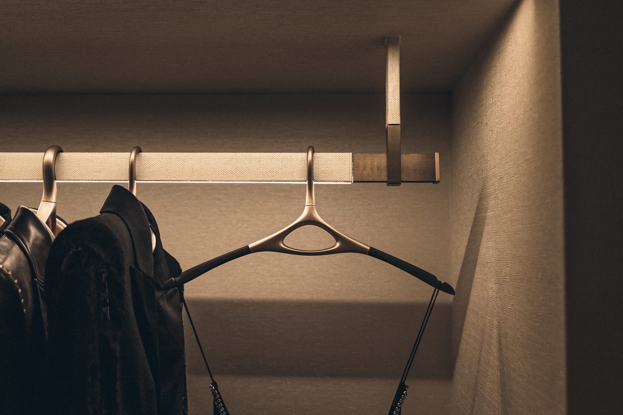 Detail of a brass wardrobe rail wrapped in linen, holding sculptural hangers and black garments under warm concealed lighting.