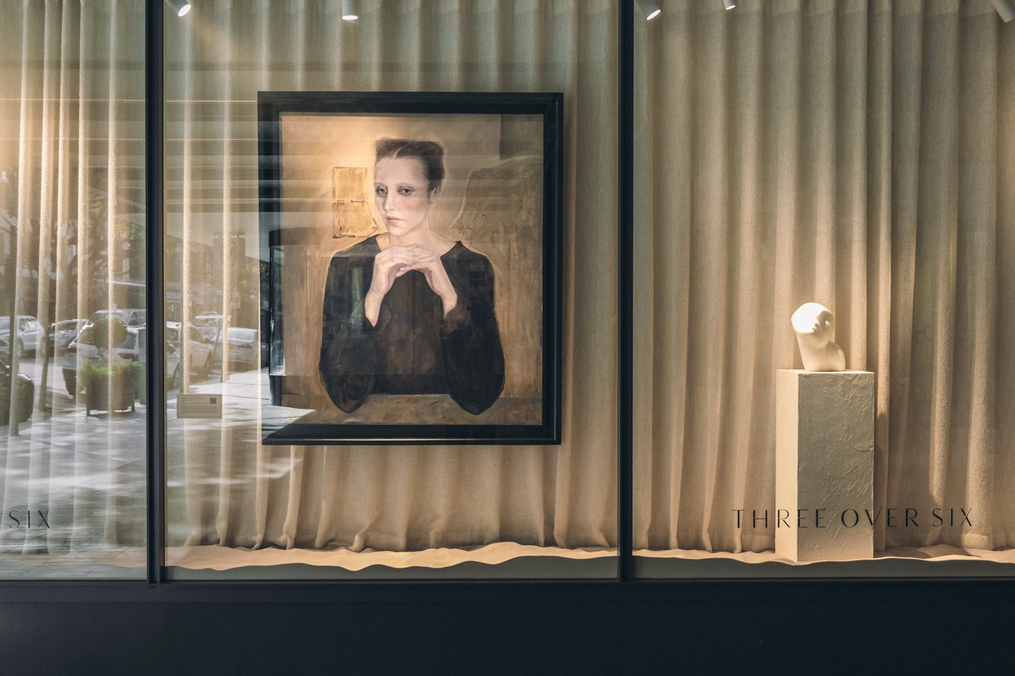 Boutique window display with a framed portrait painting and a pale bust on a plinth behind linen drapery and Three Over Six signage.
