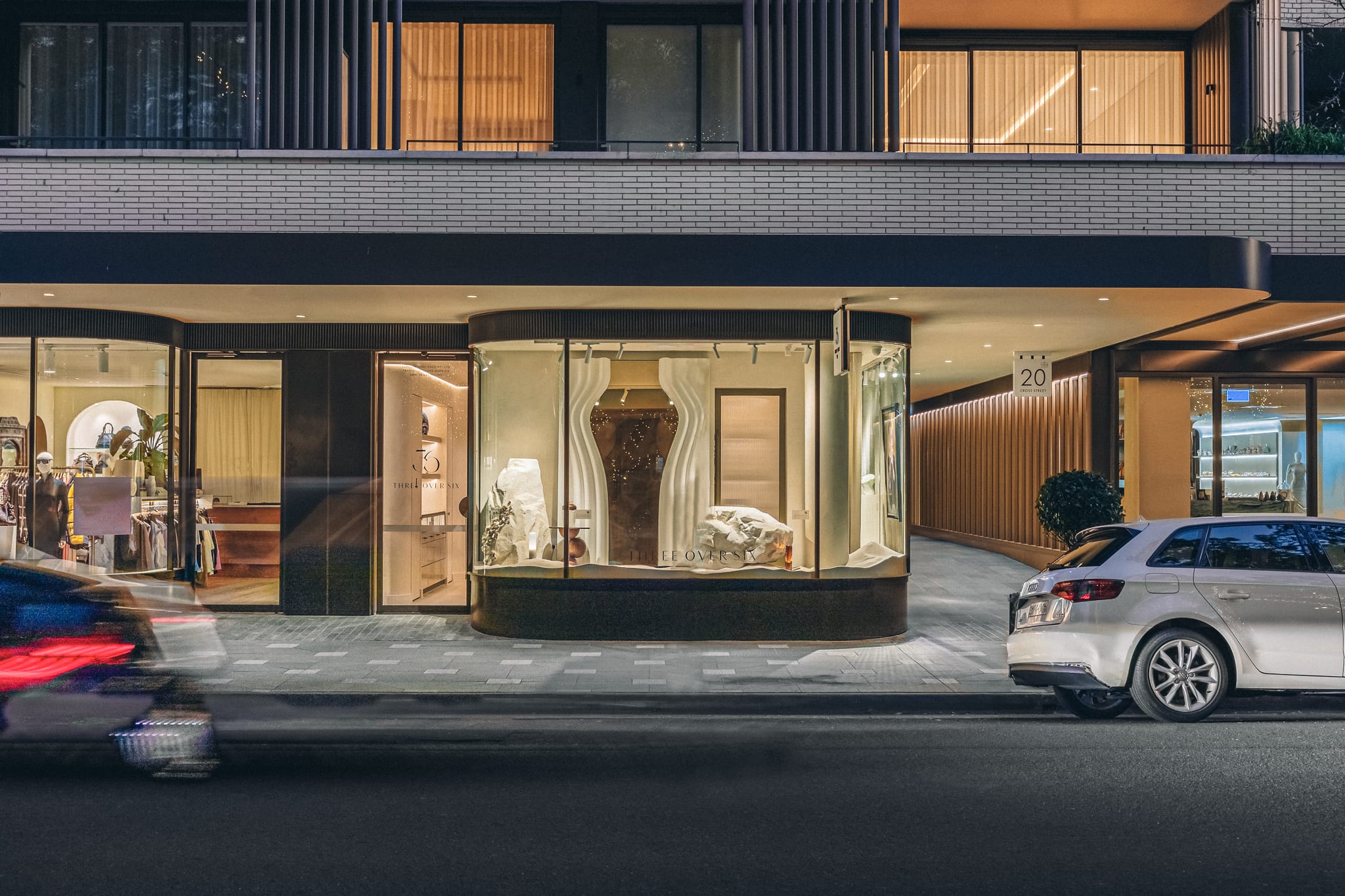 Night street view of the curved glass Double Bay shopfront lit from within, with Three Over Six signage and a sculptural window display.