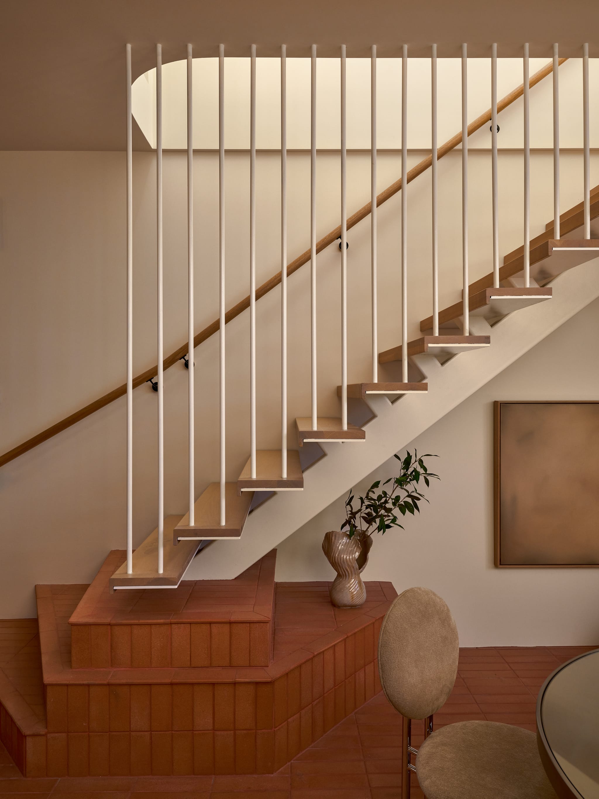 Sculptural timber staircase with slender white balusters rising from a terracotta-tiled plinth, a ceramic vase and framed artwork alongside.