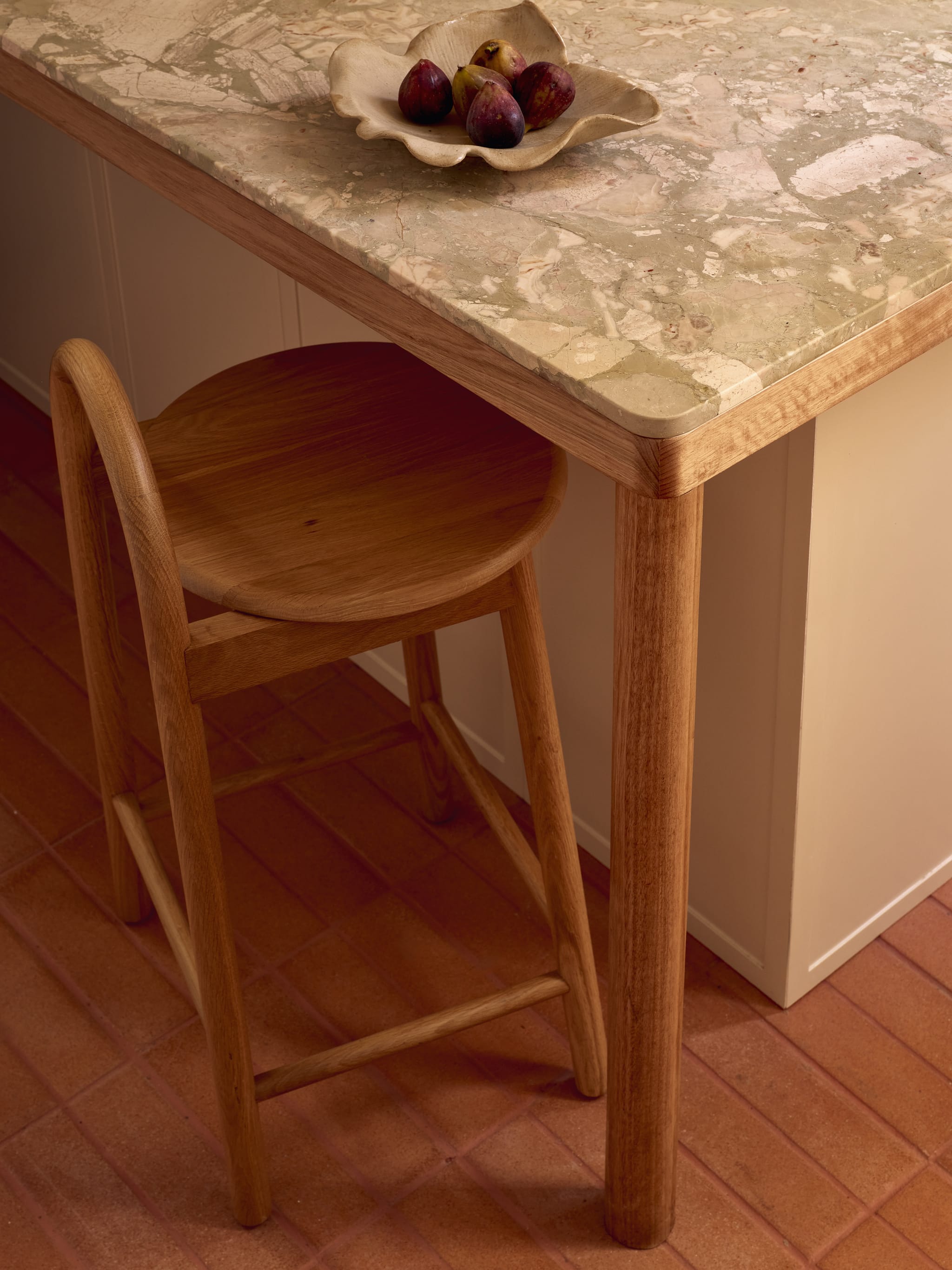 Detail of the kitchen island: veined marble top with a rounded timber edge, an oak counter stool and a dish of figs above terracotta floor tiles.