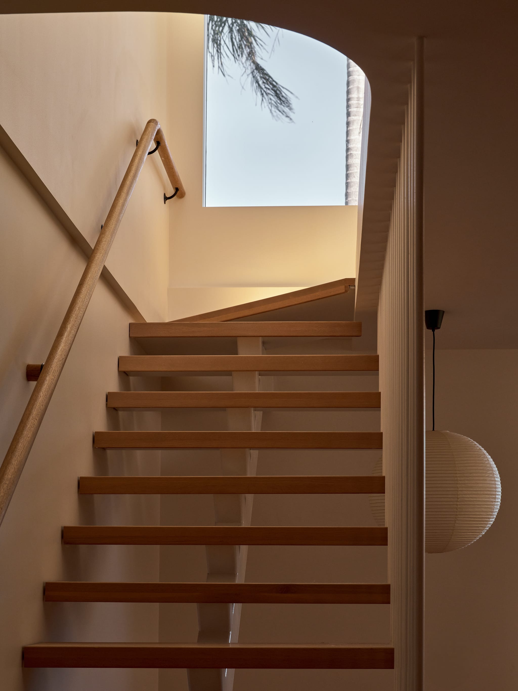 Stairwell looking up open timber treads to a skylight framing a palm, a ribbed paper lantern glowing against warm plaster walls.