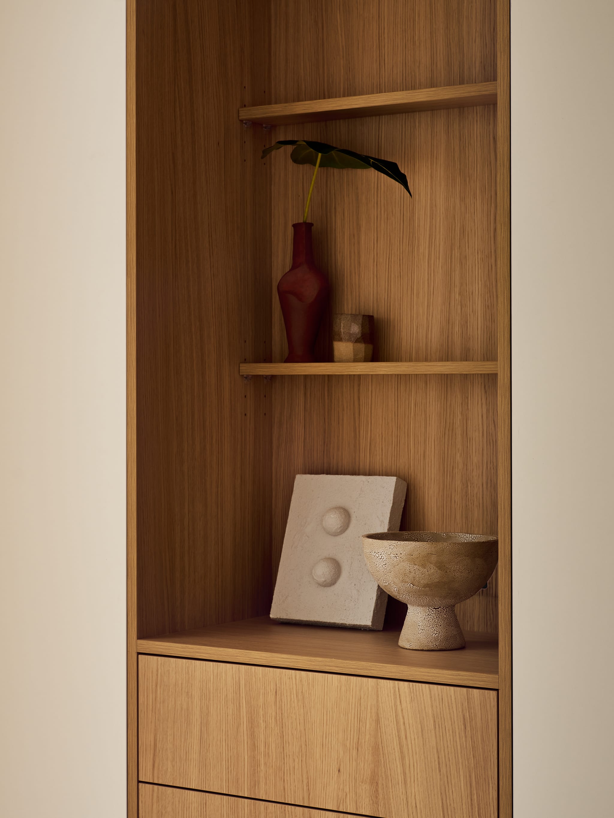 Built-in oak shelving styled with a rust-red vase, a domed plaster relief and a footed ceramic bowl, with drawers set into the joinery below.