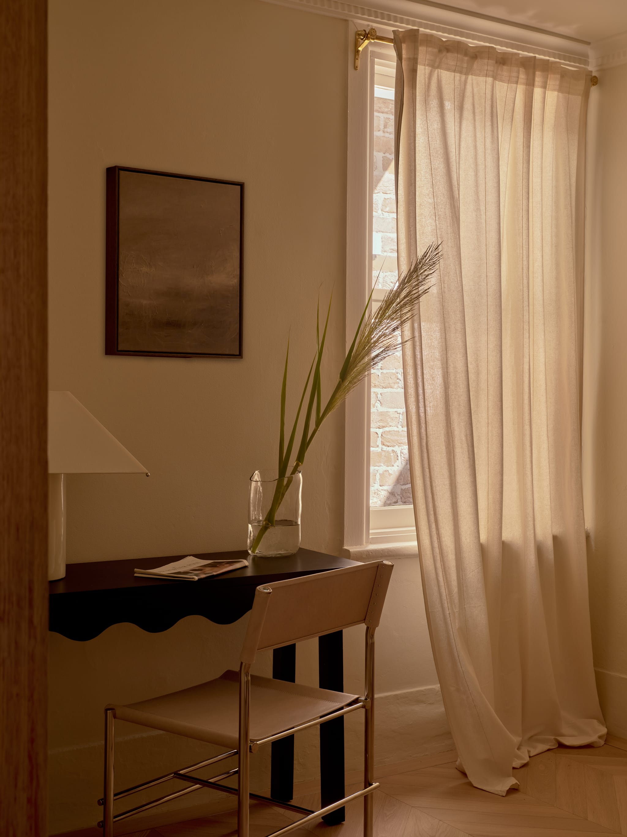 Study nook with a dark scallop-edged desk, a leather sling chair and a sheer-curtained window revealing painted brick, on herringbone timber floor.