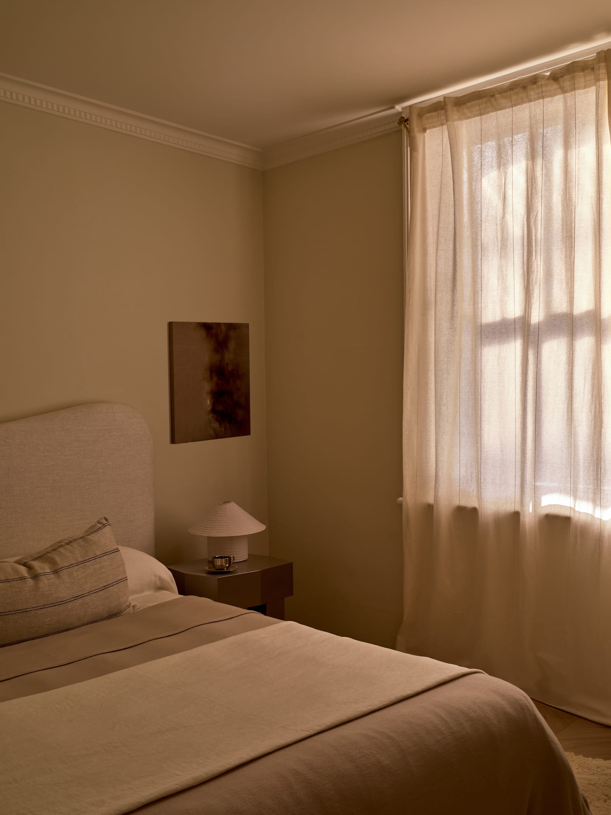 Bedroom in warm light with an upholstered headboard, a pleated-shade lamp and teacup on the bedside table, and glowing sheer curtains under a cornice.
