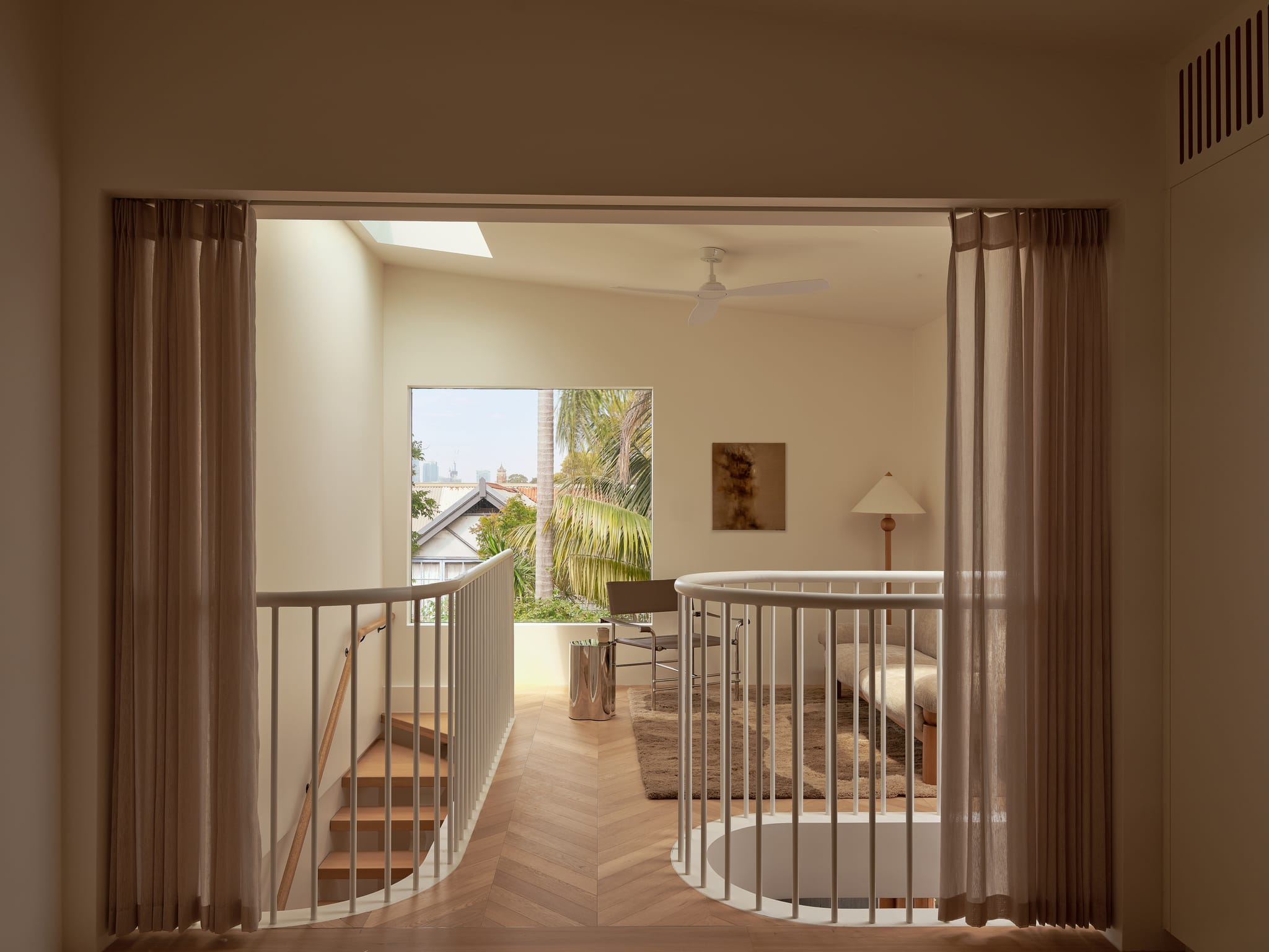 Upper living level of the Birchgrove home framed by curtains: a curved balustrade around the stair void and a window over rooftops and palms.