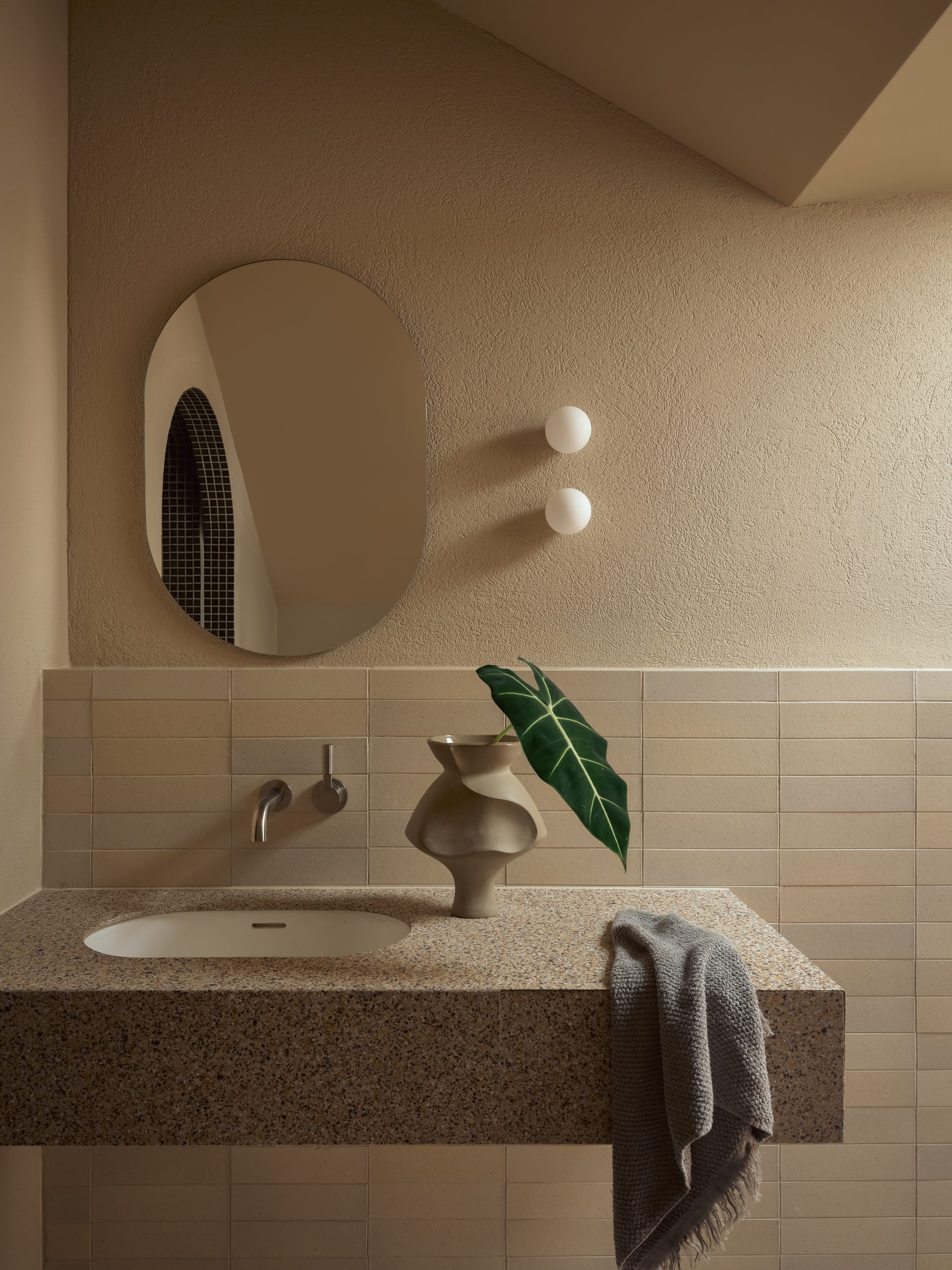 Bathroom with a terrazzo vanity, wall-mounted tap and oval mirror reflecting an arched mosaic shower, spherical wall lights on textured plaster.