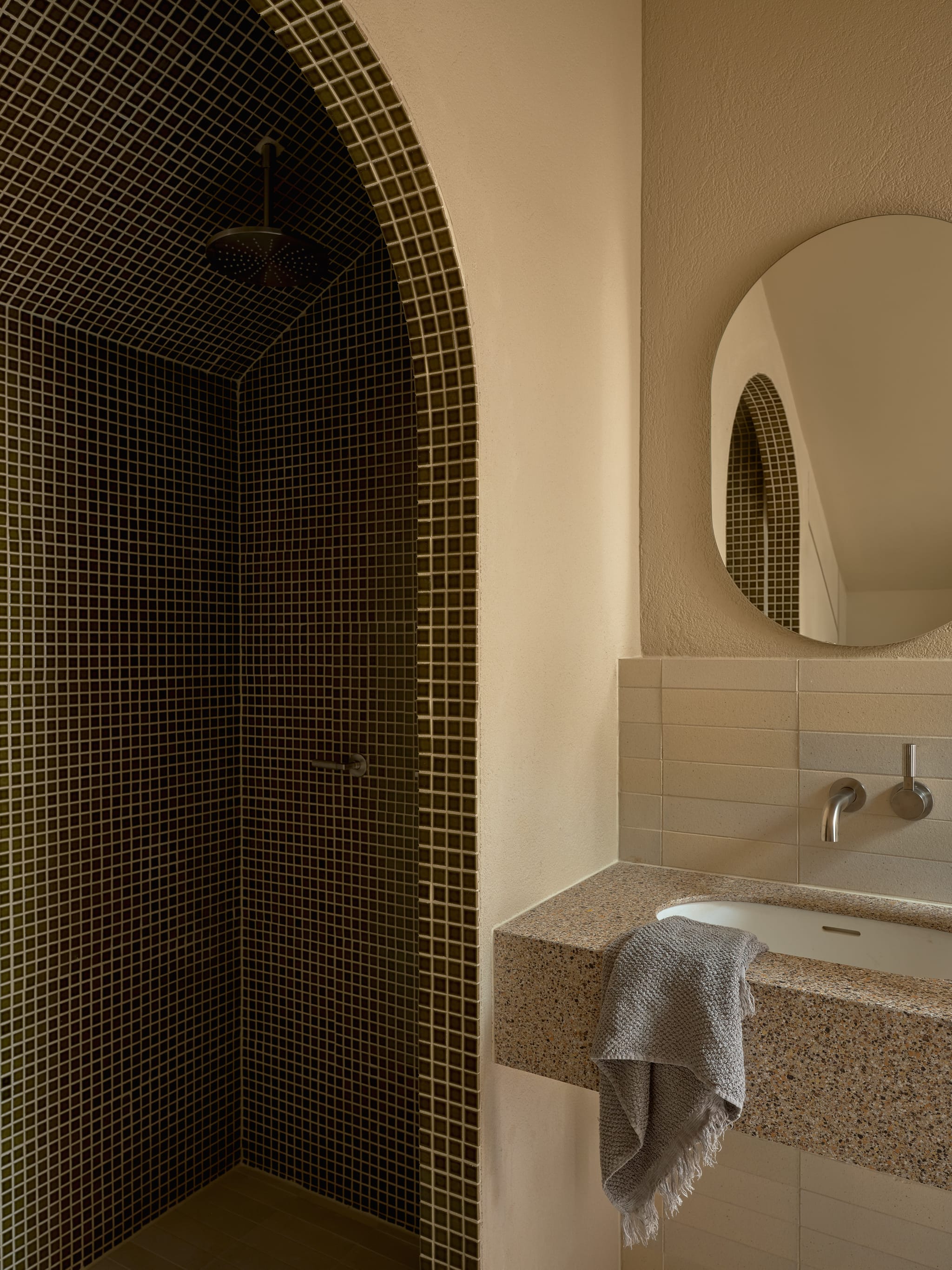 Arched shower recess clad in dark mosaic tiles with a rain head, beside a terrazzo vanity, oval mirror and a fringed grey towel.