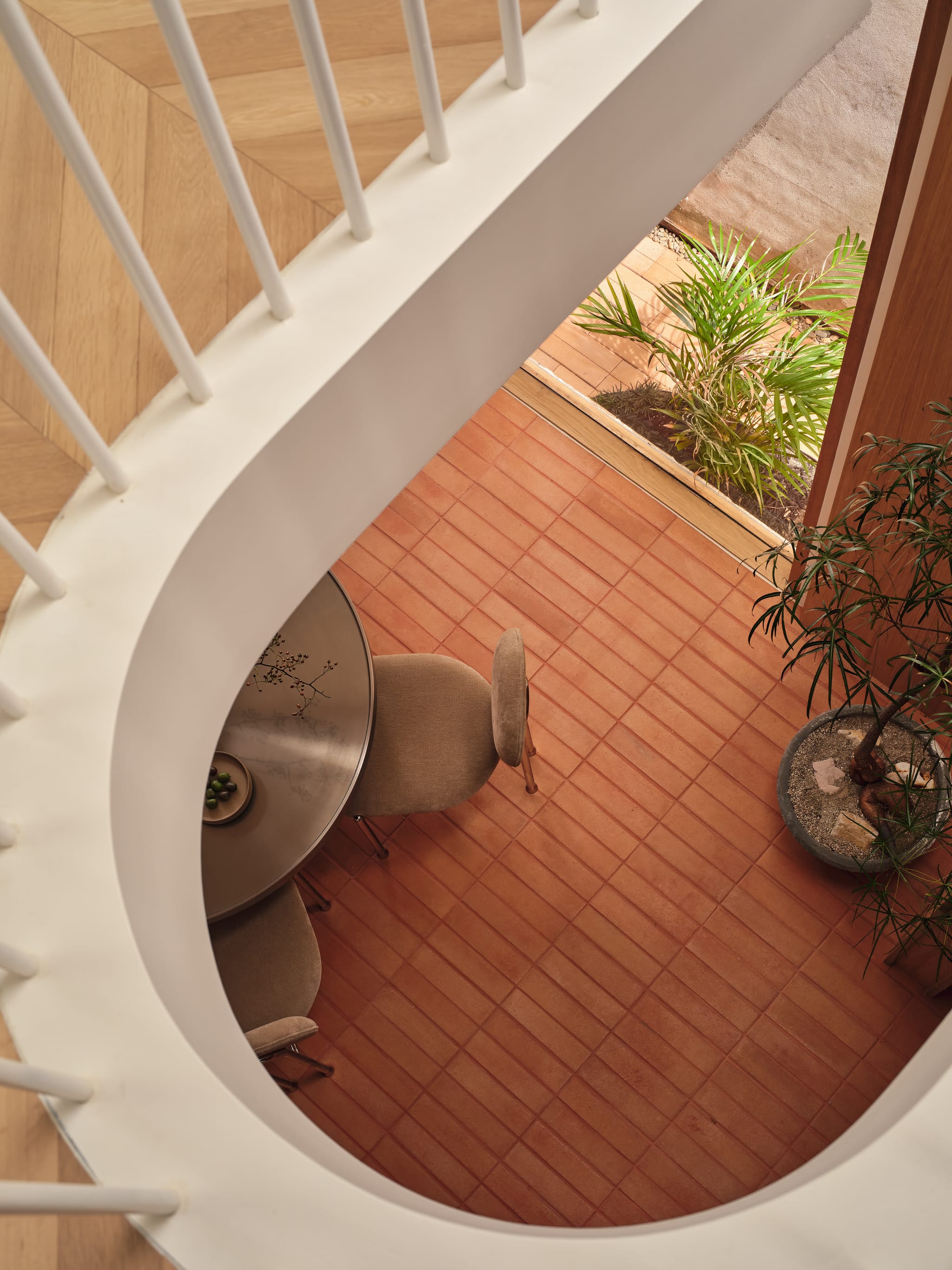 Looking down through the oval stair void to terracotta tiles, a round dining table with upholstered chairs and potted greenery in the pavilion below.