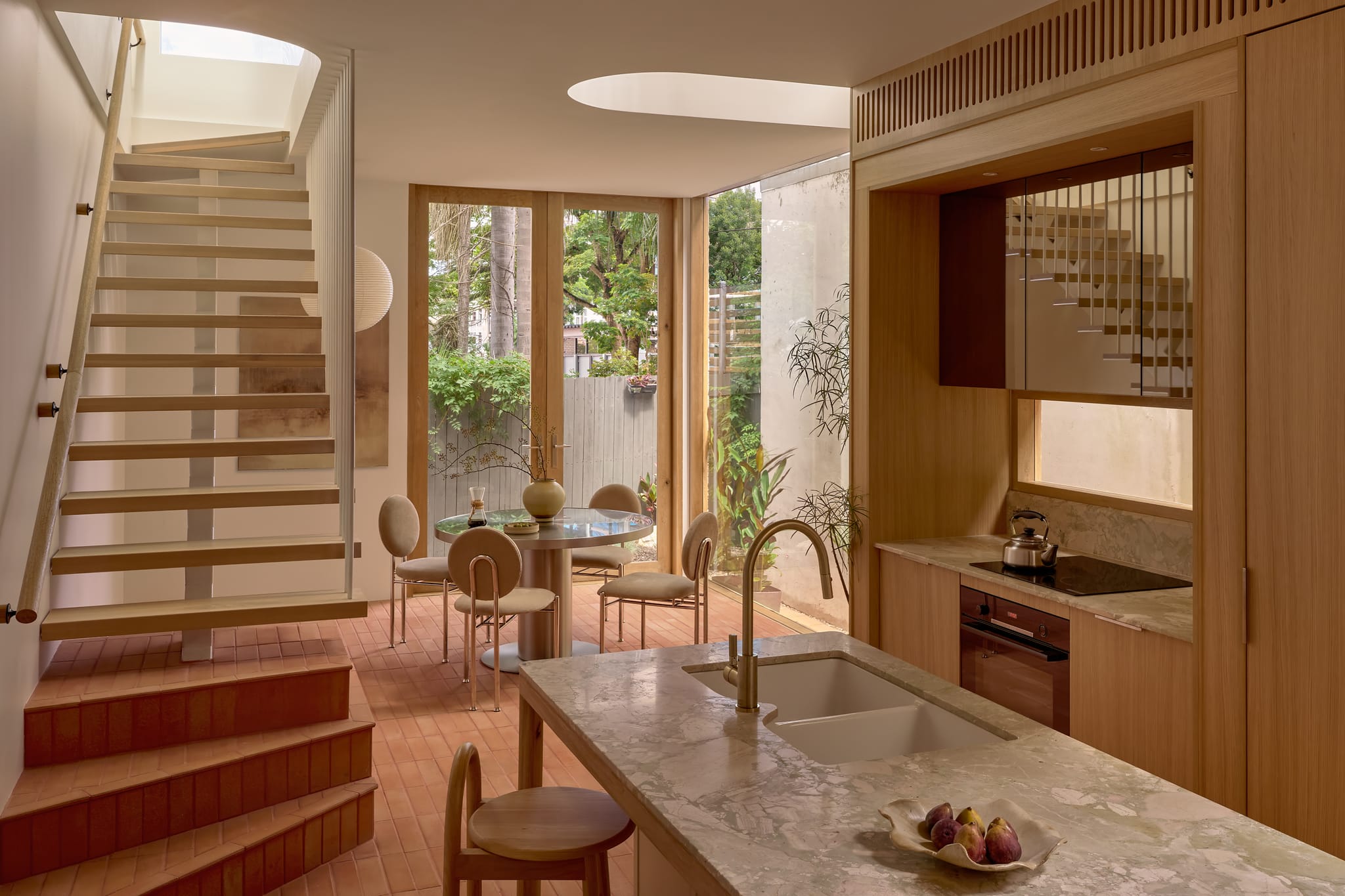 Rear pavilion kitchen with a marble island and double sink, open-tread oak stair, terracotta floor tiles and glass doors to a planted courtyard.
