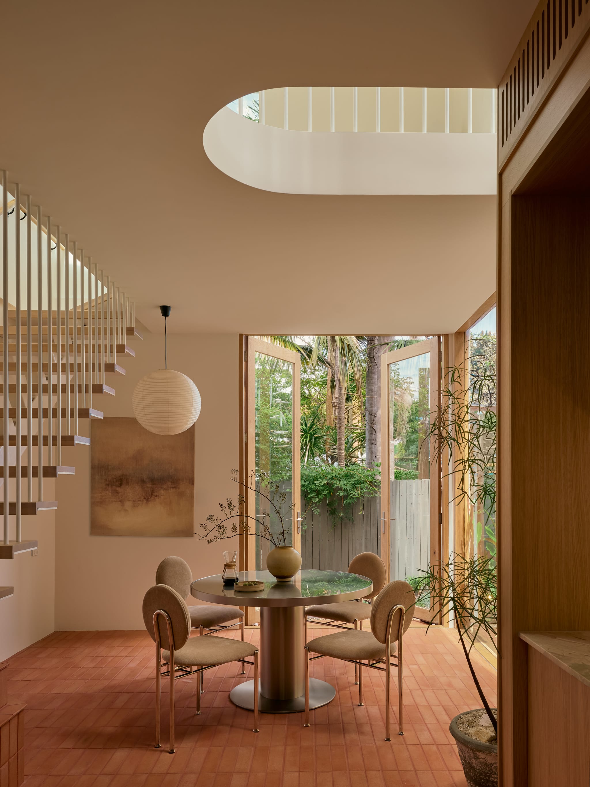 Rear pavilion dining under an oval skylight void: a floating timber stair, paper lantern, round table and French doors to a palm-filled garden.