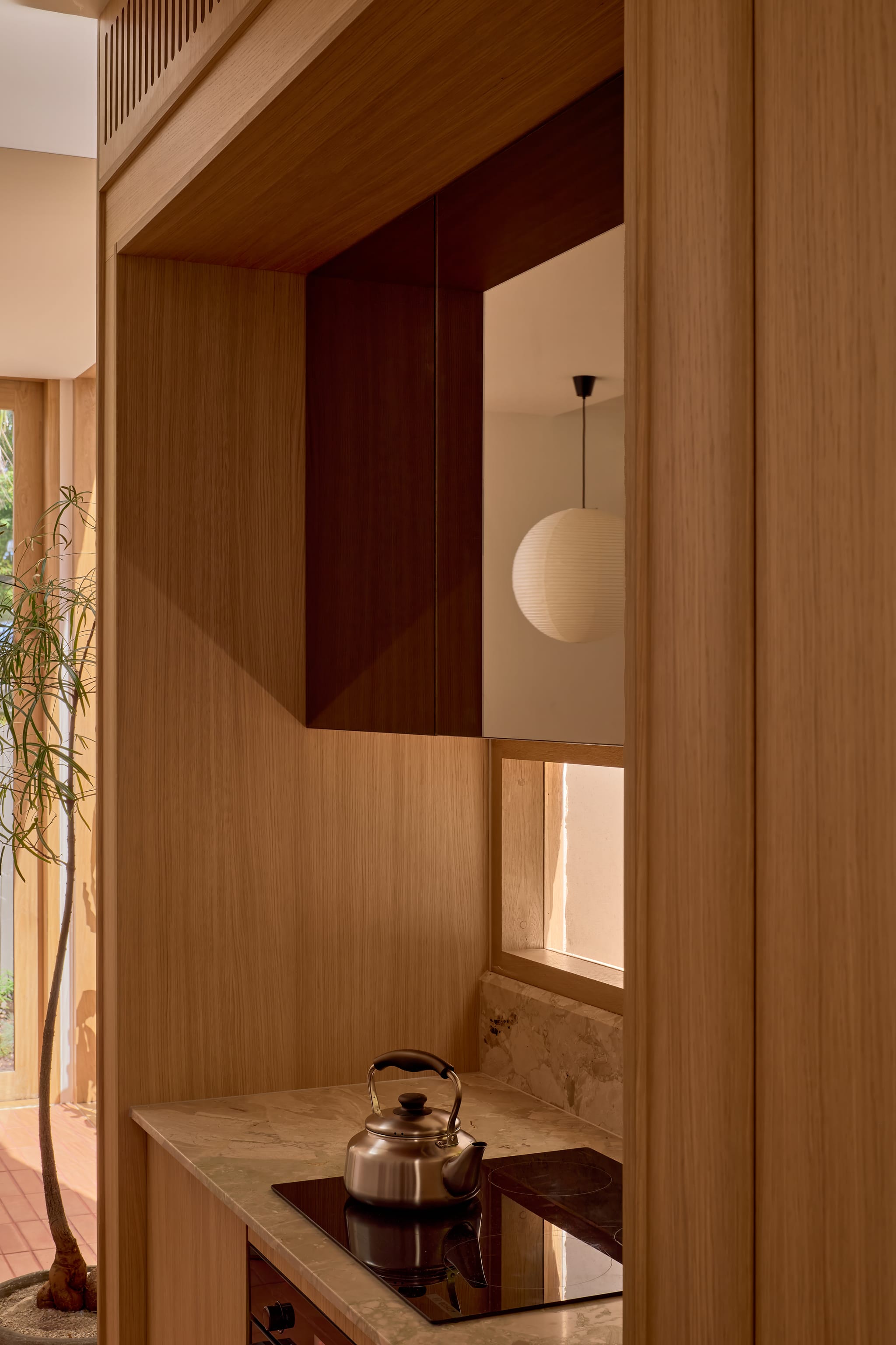 Kitchen framed in oak joinery, a kettle on the cooktop, marble benchtop and splashback, and a tinted mirror reflecting a paper lantern overhead.