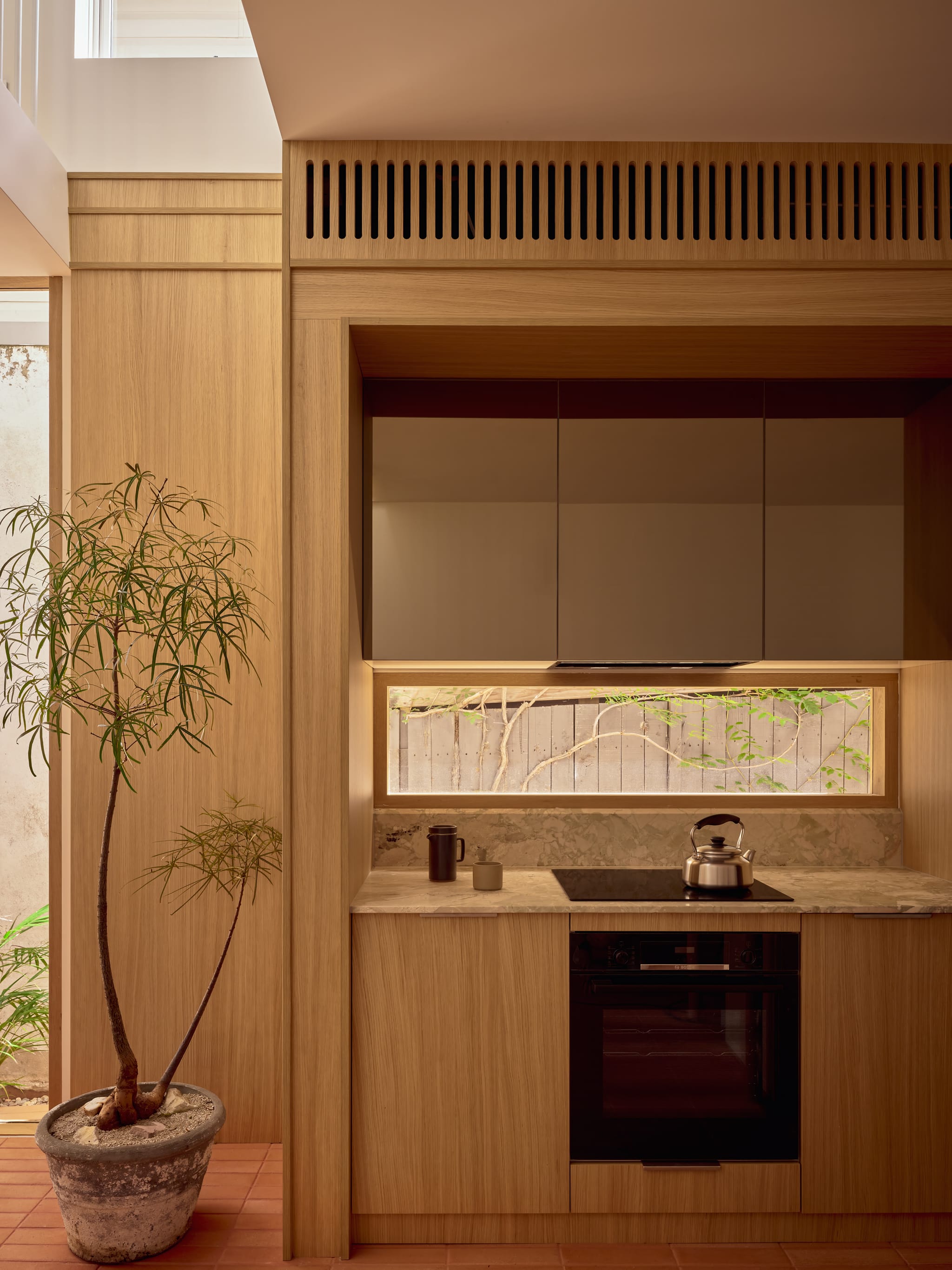 Compact oak kitchen alcove with marble benchtop, induction cooktop and a low horizontal window to the courtyard, a potted plant on terracotta tiles.