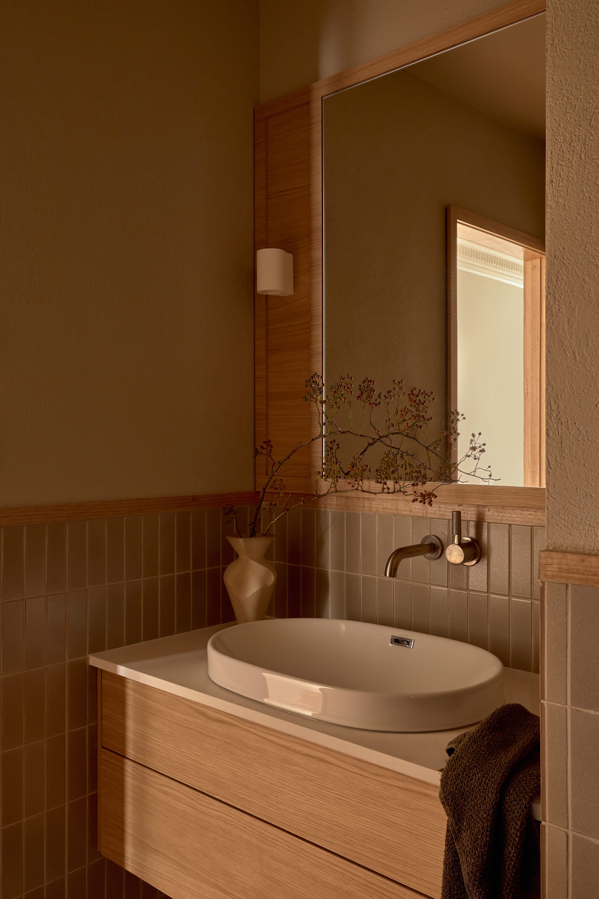 Powder room with sage stack-bonded tiles, an oak vanity and drop-in basin, a berried branch in a ceramic vase and a mirror reflecting the doorway.