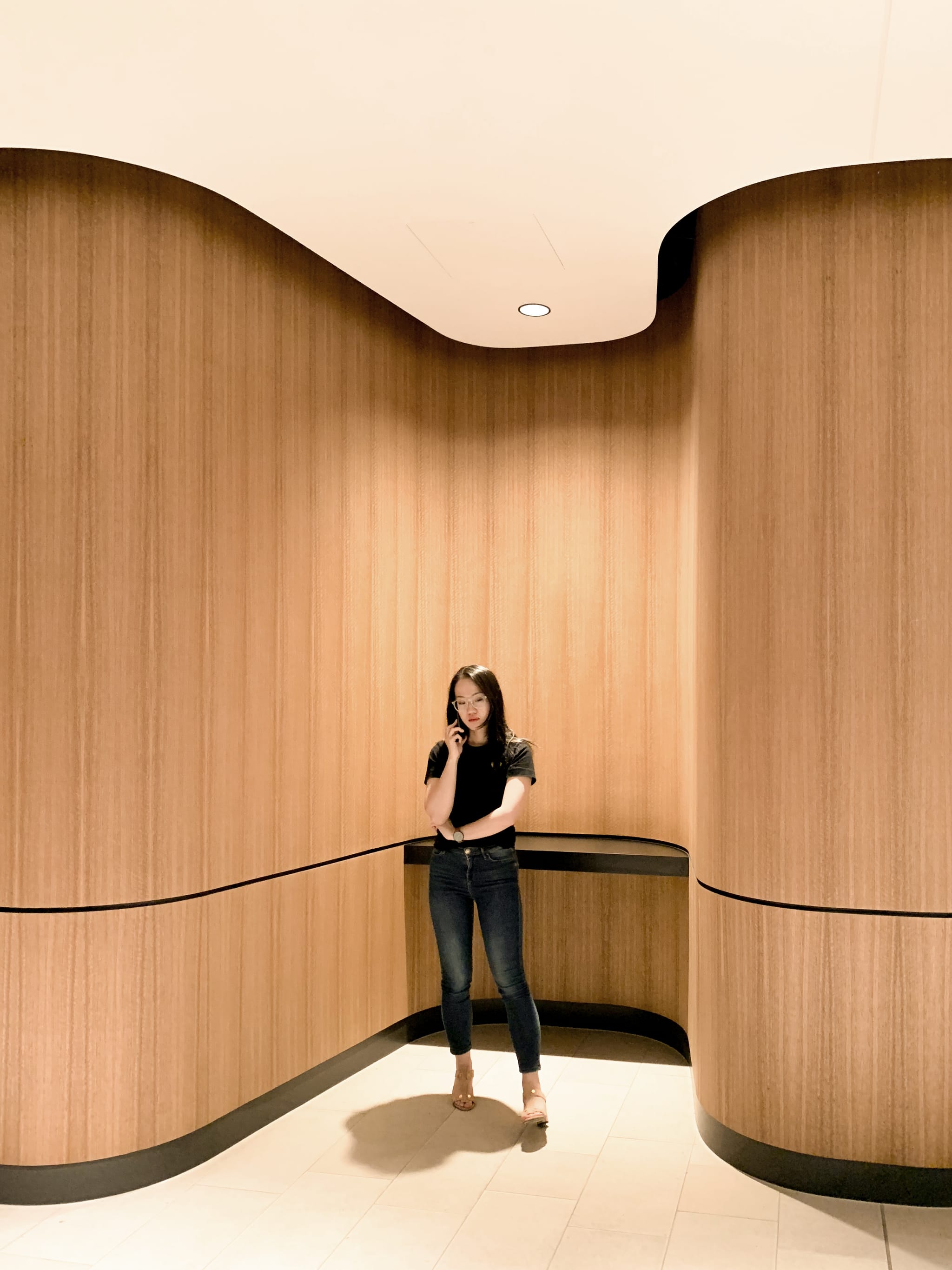 A woman on her phone stands against a sculpted curved timber-veneer wall in the workplace lobby, cream tiled floor underfoot