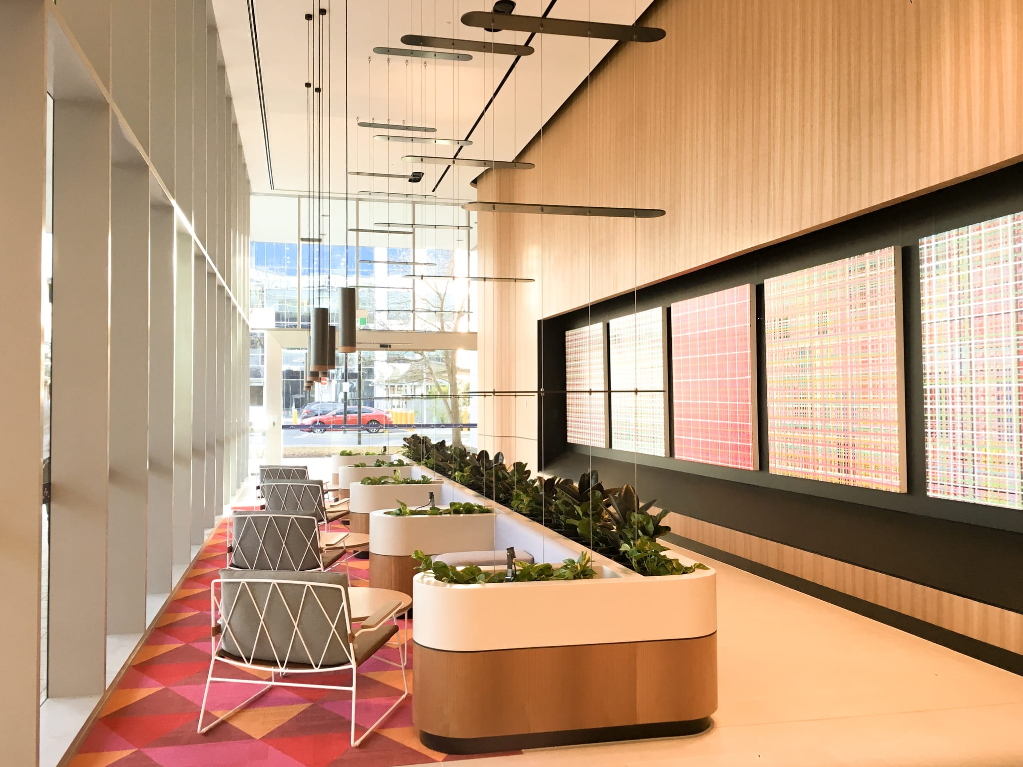 Lobby seating runs beside backlit red woven-grid art panels and planters, with glazing to the street beyond, at Macquarie Park