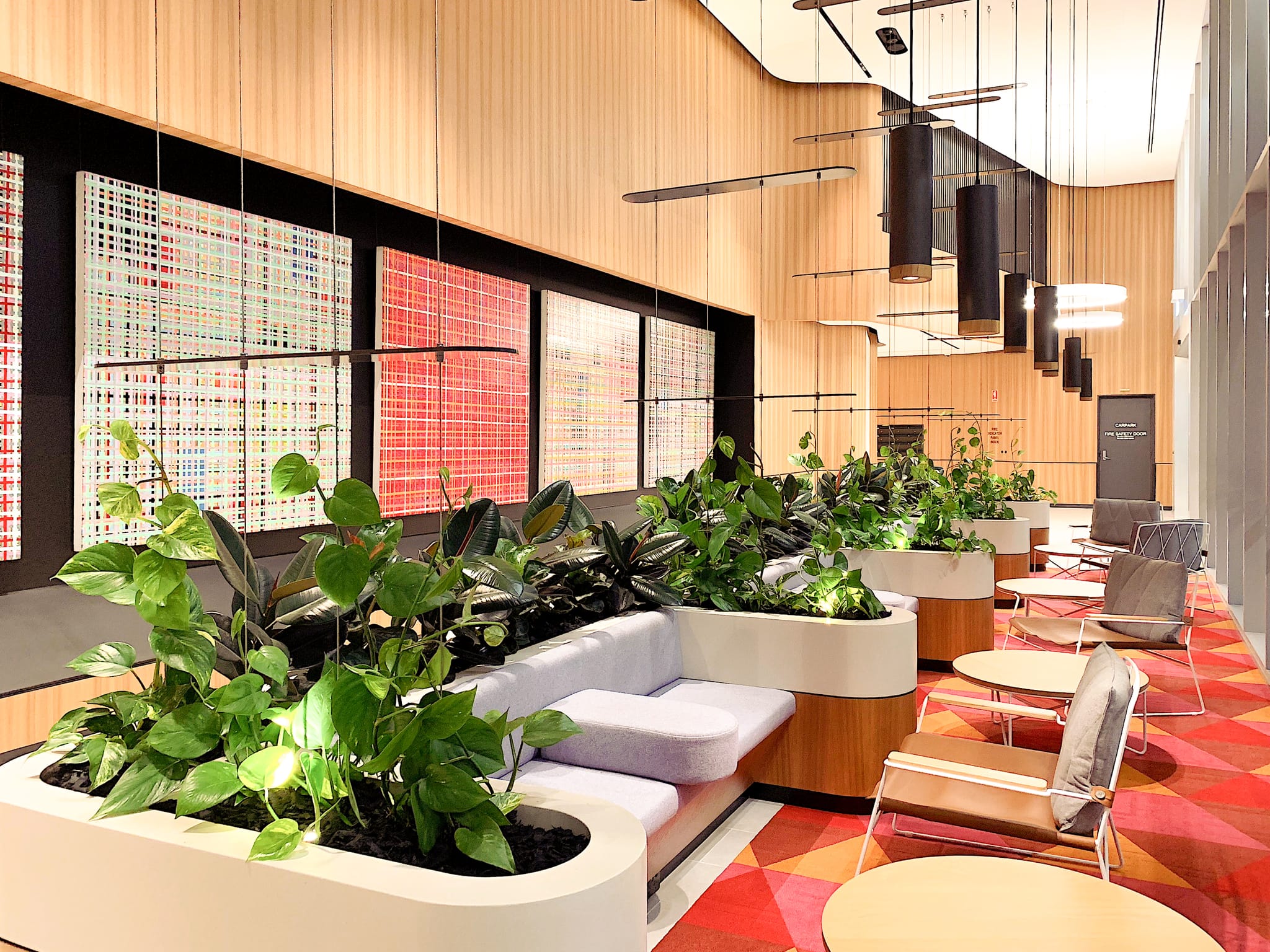 Planted bench seating and leather armchairs line colourful backlit artwork panels above patterned carpet in the commercial interior