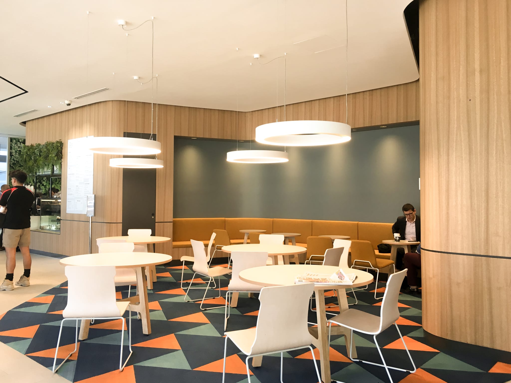 Cafe seating with round white tables, an orange banquette and circular pendant lights beside a green living wall in the workplace lobby