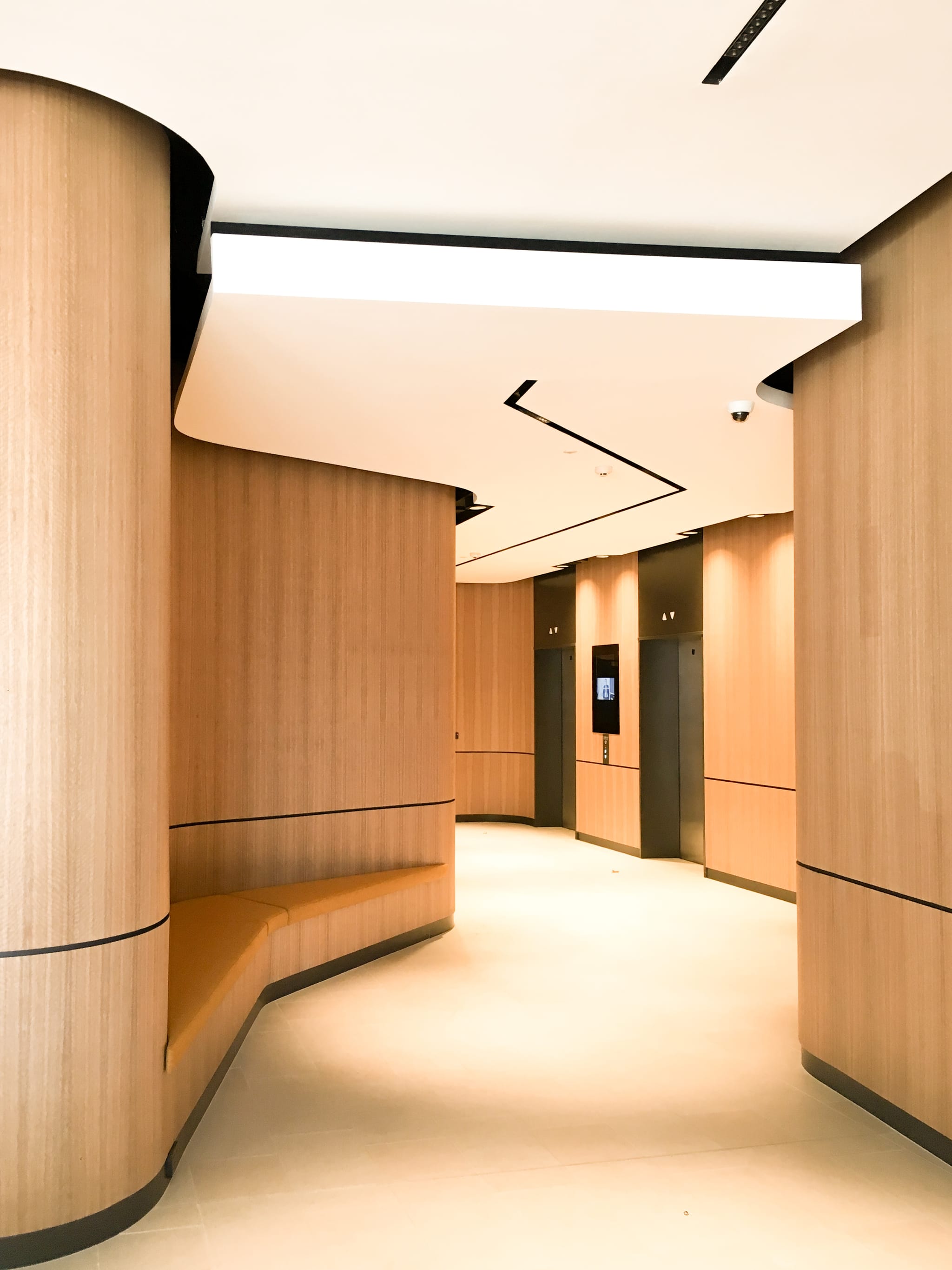 Lift lobby framed by curved timber-veneer walls with a built-in bench nook and warm downlights in the Macquarie Park commercial interior