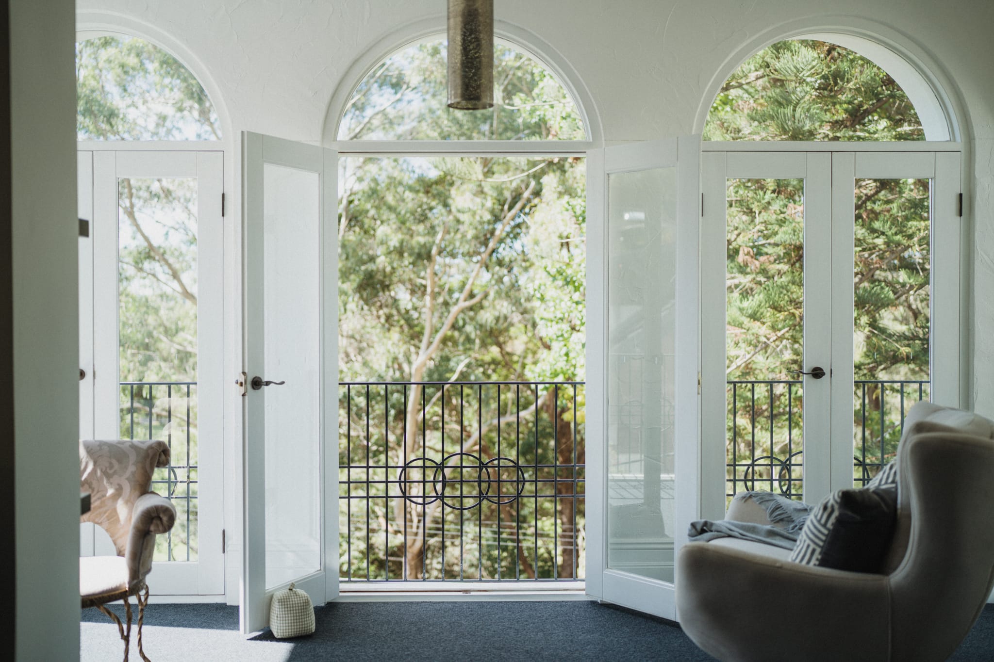 Arched French doors open to a wrought iron Juliet balcony framed by treetops, with an upholstered armchair and curved sofa in a light-filled sitting room.