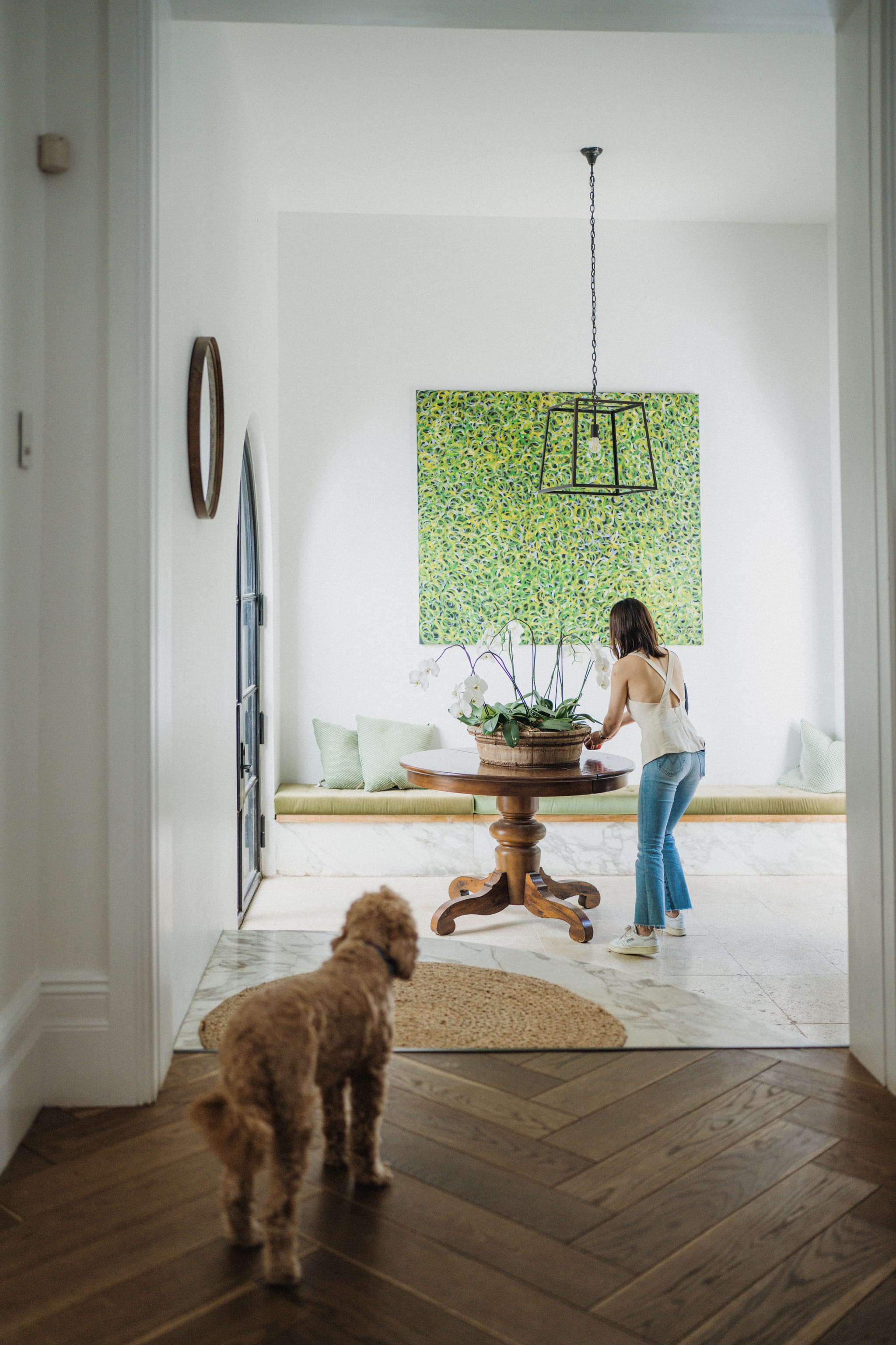 A woman arranges white orchids on a round timber pedestal table as an apricot dog looks on, beneath green abstract art on herringbone floors.