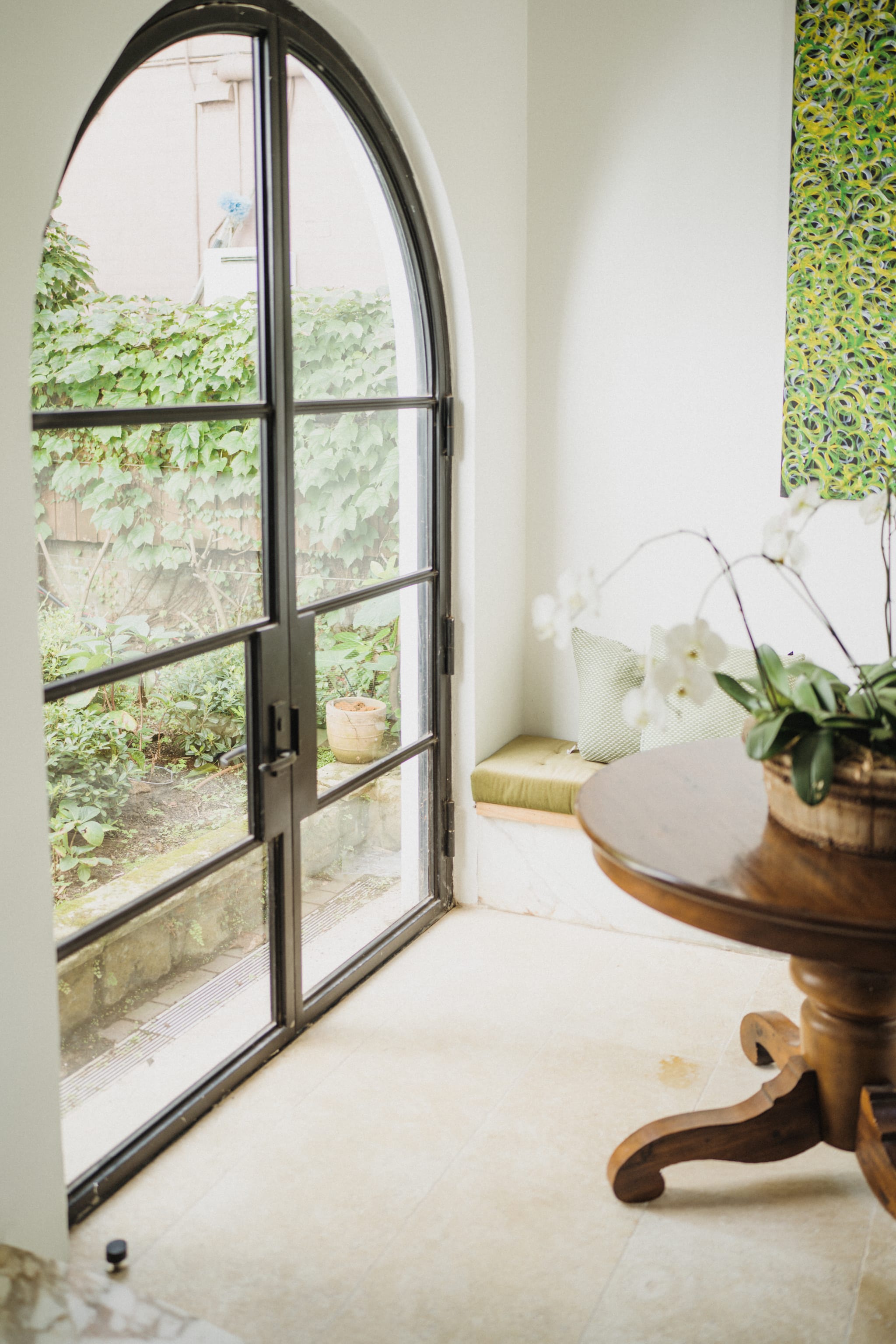Arched steel-framed glass door opens to a leafy garden beside a cushioned window seat and a timber table set with white orchids, on pale stone floors.