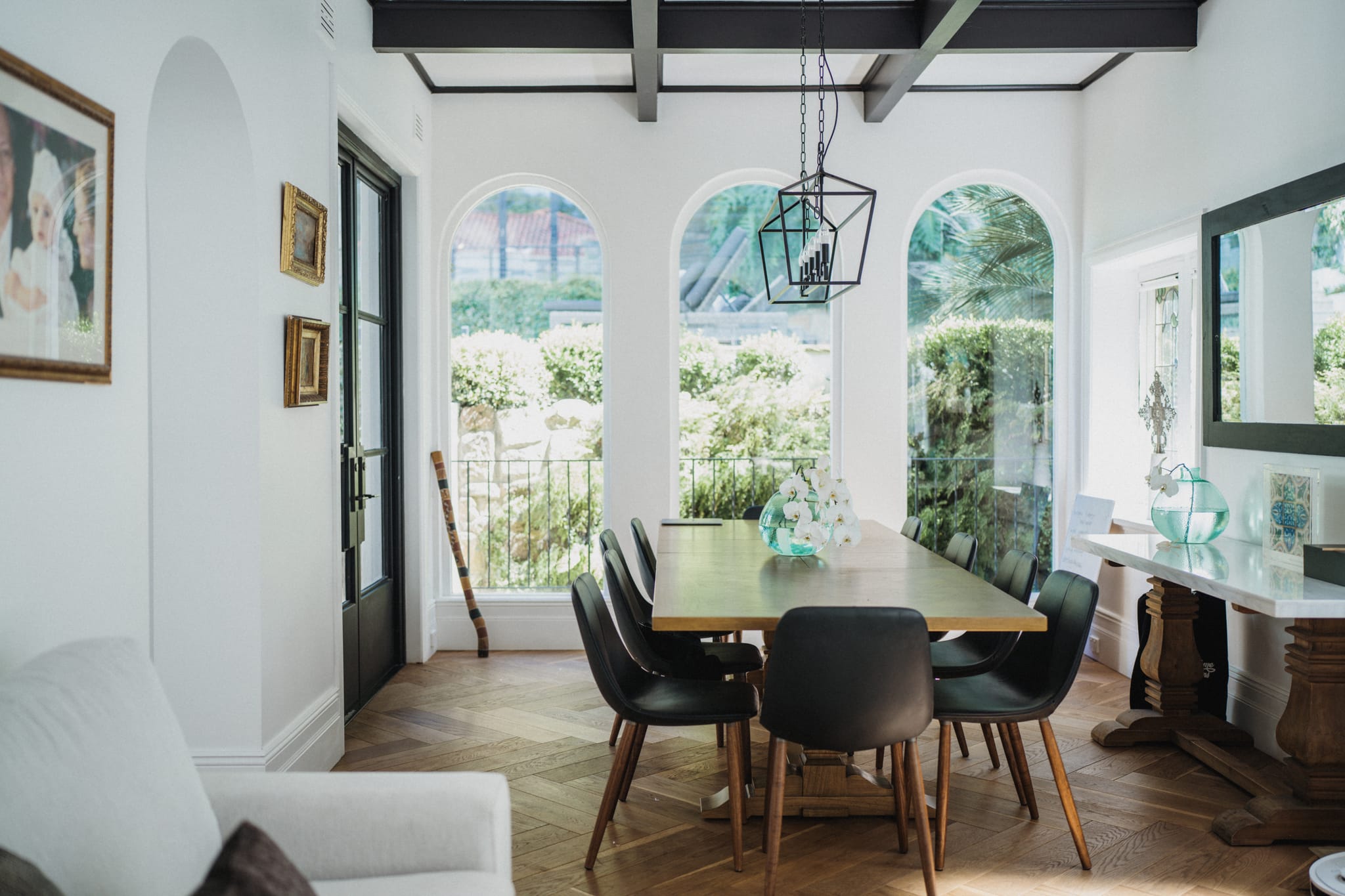Dining room with a long timber table and black leather chairs by arched garden windows, below dark exposed ceiling beams on oak herringbone parquet.