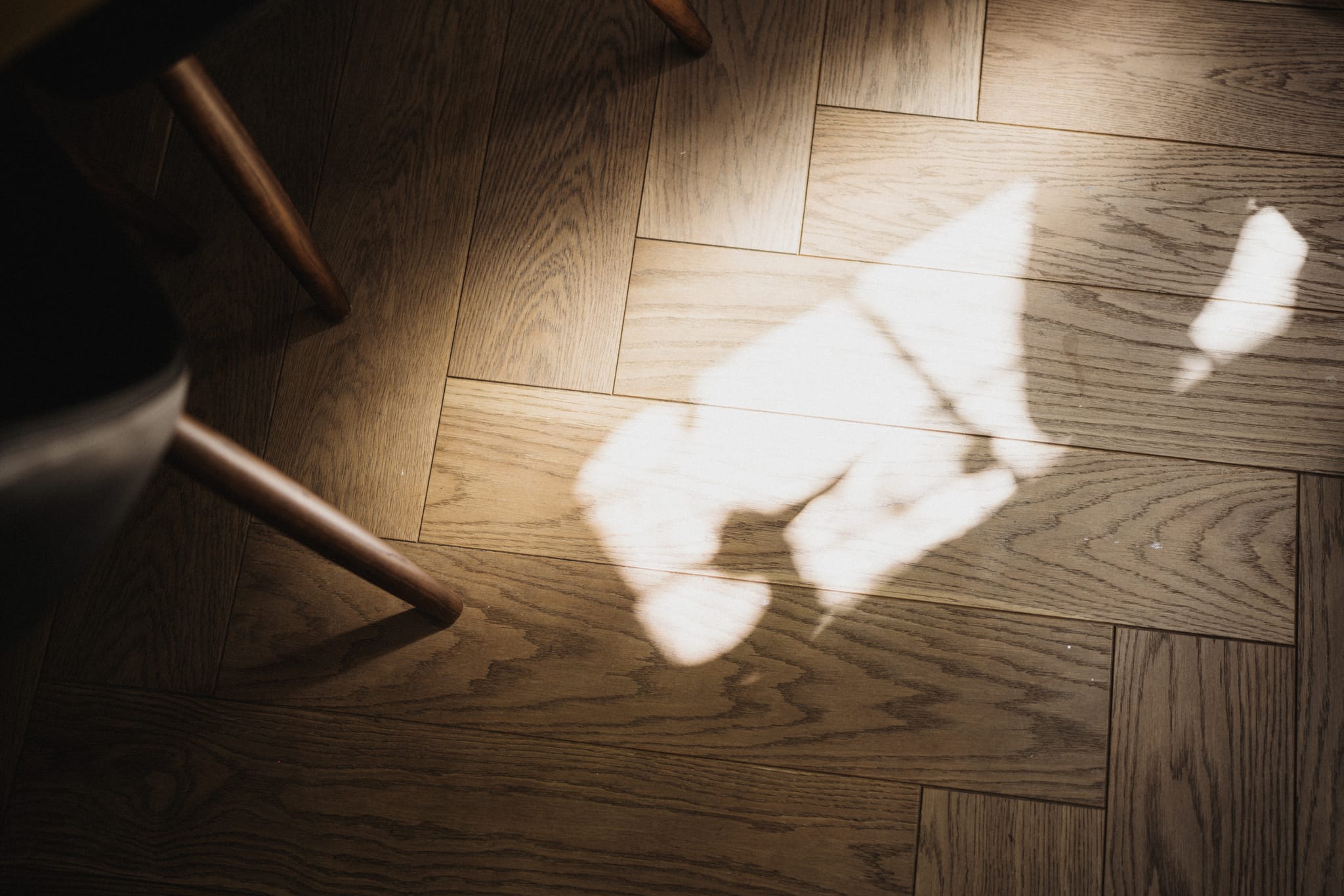Sunlight casts a windowpane pattern across warm oak herringbone parquet, with timber chair legs at the edge of the family home's dining space.