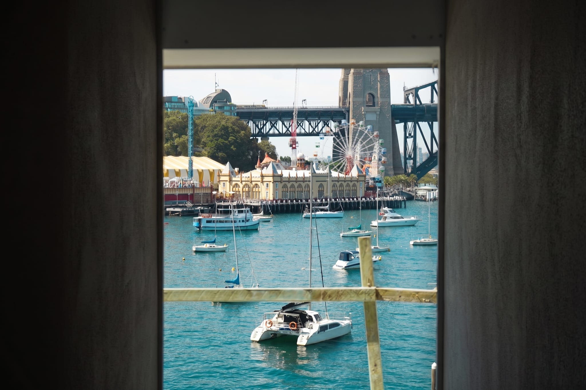 Harbourside outlook through a narrow window: boats moored in Lavender Bay below Luna Park, the ferris wheel and a Sydney Harbour Bridge pylon